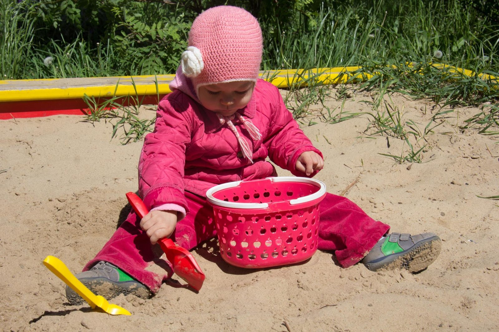 A young child wearing a pink knit hat and pink jacket sits in sand playing with a red perforated basket and sand toys including a yellow shovel.