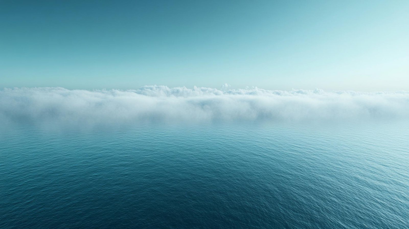 A view of calm turquoise ocean water in the foreground with a layer of white clouds hovering above the horizon and a clear blue sky above.