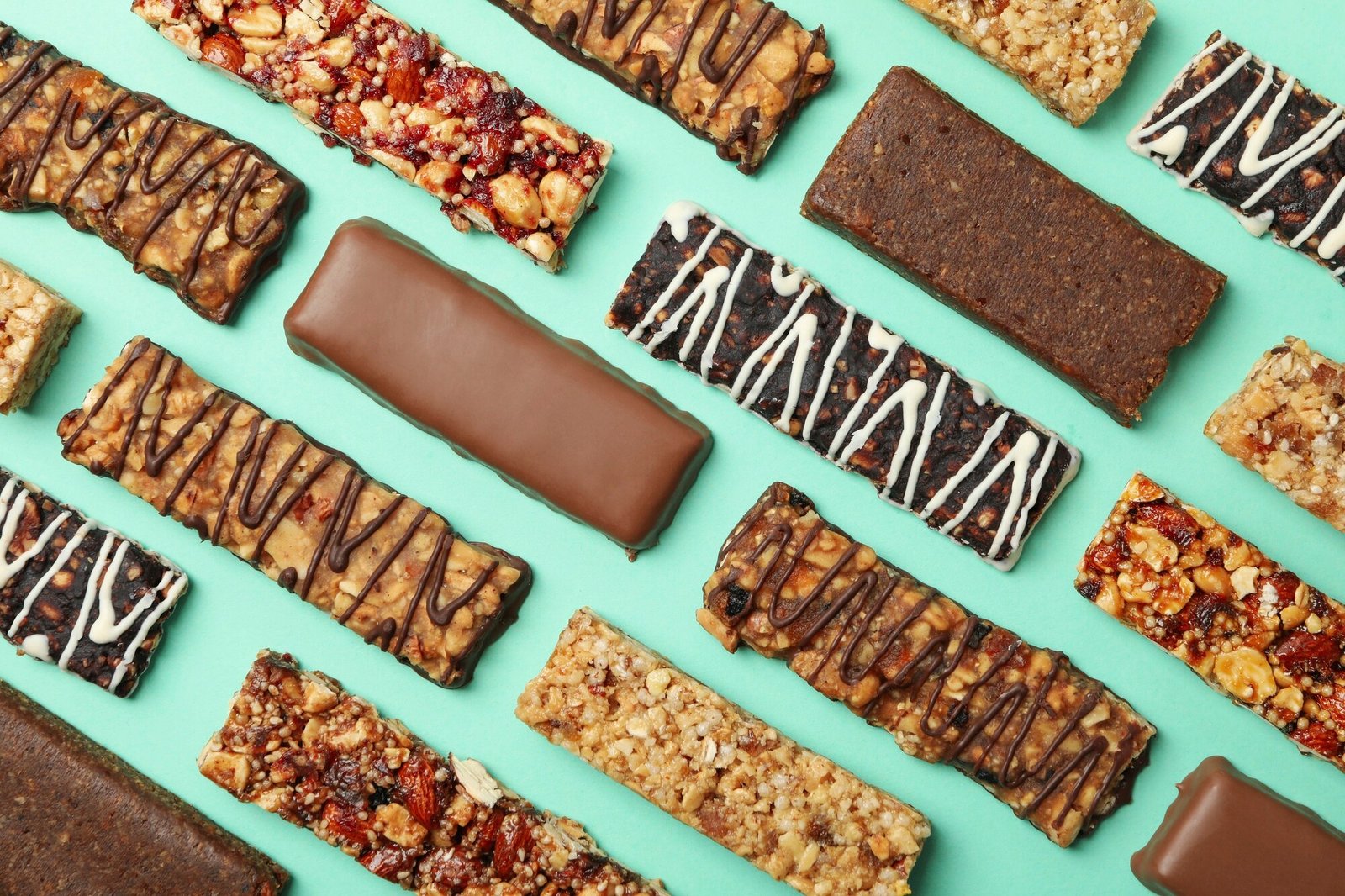 An overhead view of various granola and protein bars arranged on a light blue surface, including bars with chocolate drizzle, white icing drizzle, chocolate coating, and visible nuts and dried fruit.