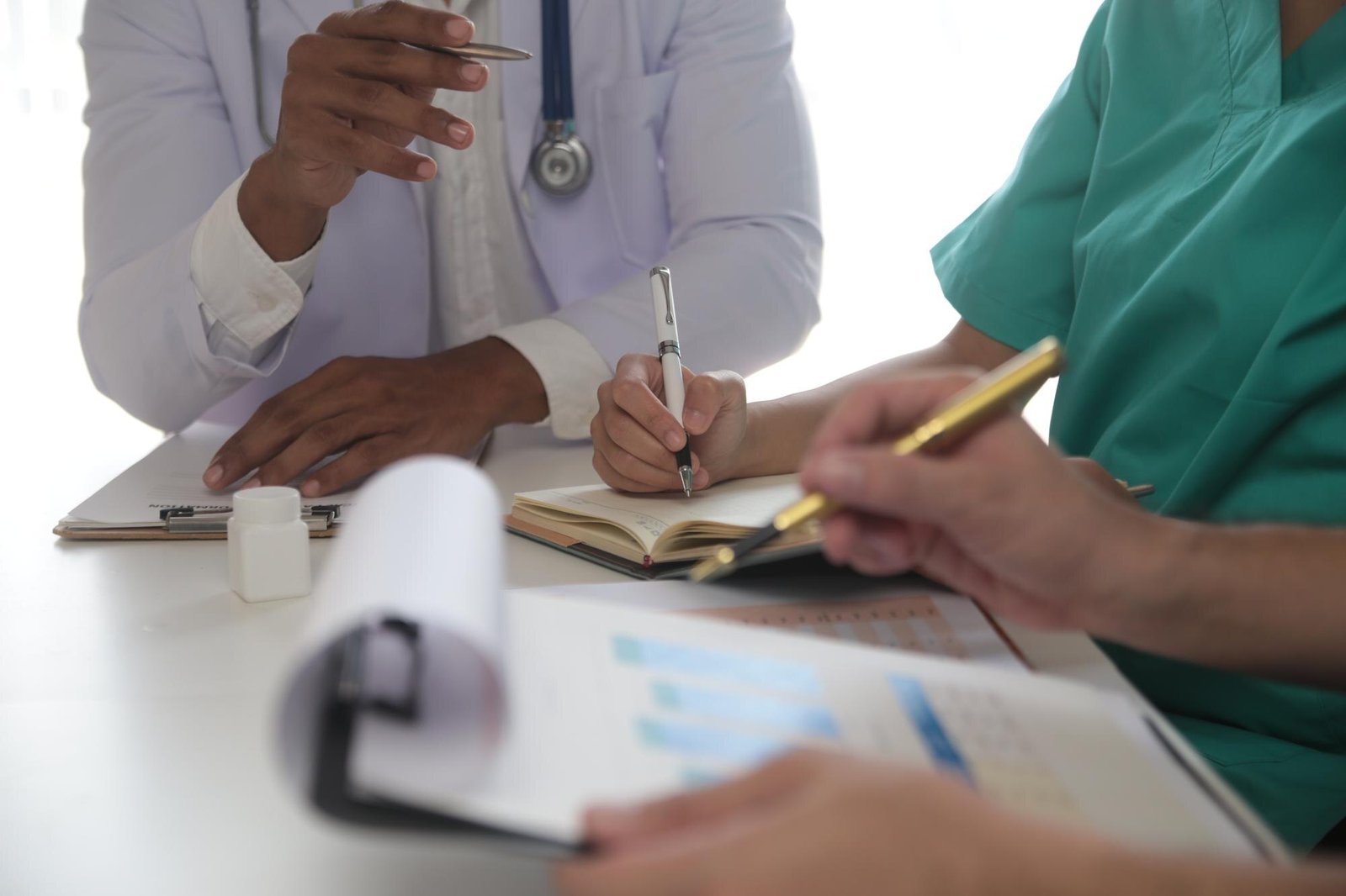 Medical professionals in white coat and green scrubs sit at a table writing in notebooks with pens, with a coffee cup and papers visible on the desk.