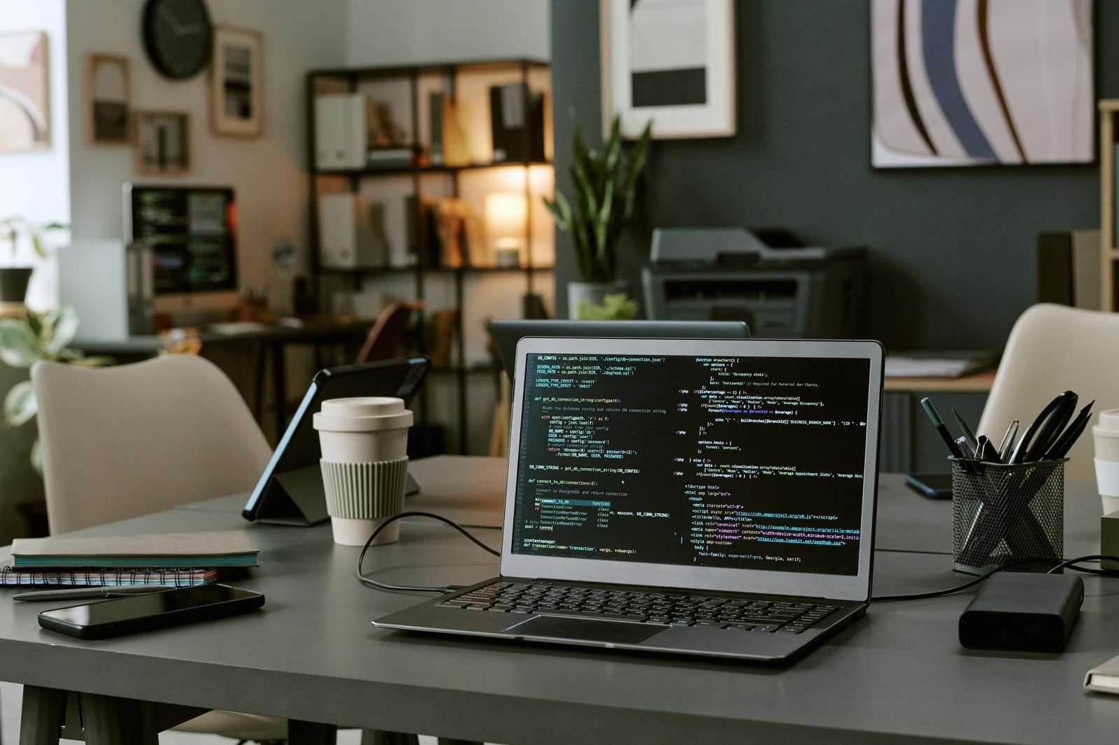 A laptop displaying code on its screen sits on a desk alongside a coffee cup, tablet, notebooks, pen holder, and external hard drive, with a home office environment visible in the background.