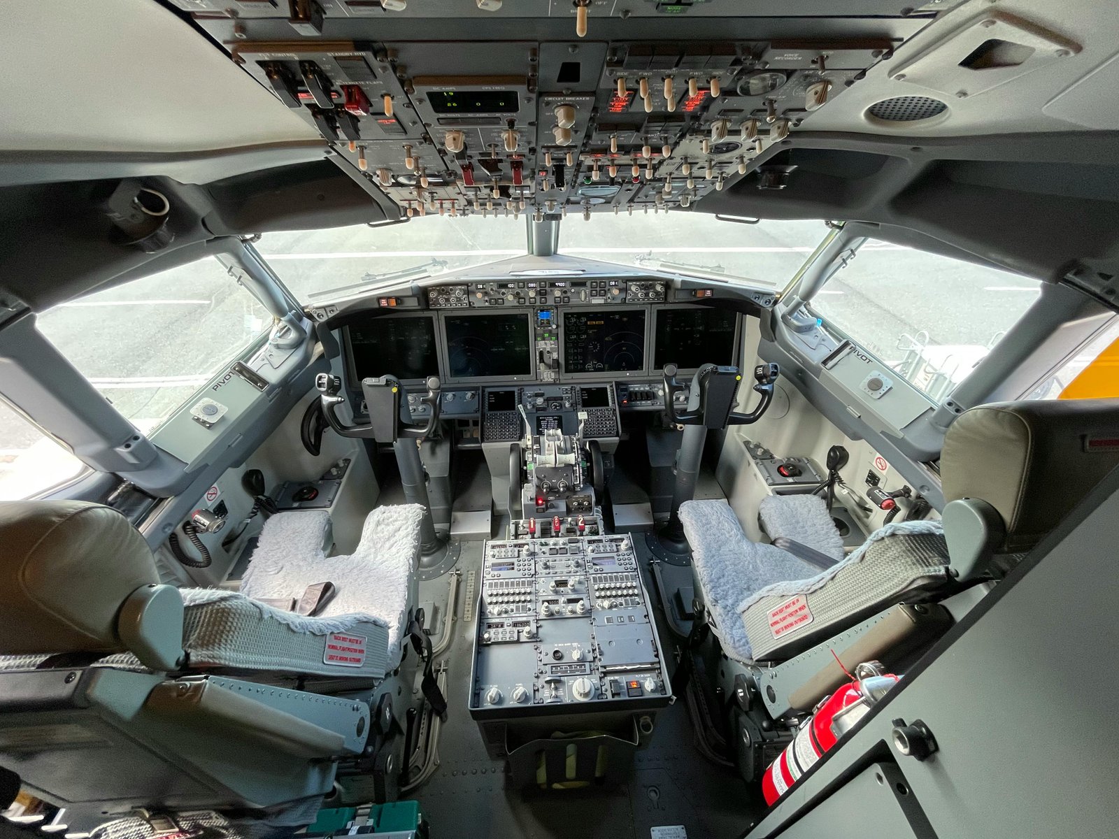 View from inside an aircraft cockpit showing the flight deck with two pilot seats, multiple instrument panels, screens, and control systems in a commercial aviation setting.