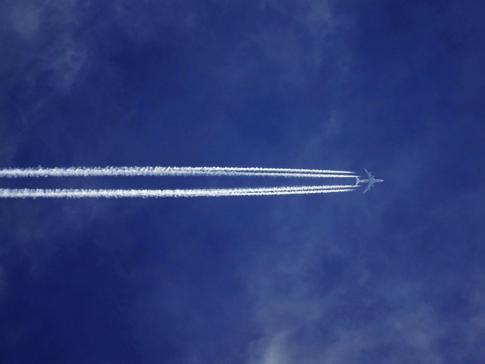 A high-altitude aircraft contrail stretches across a deep blue sky, leaving parallel lines of white condensation clouds in its wake.