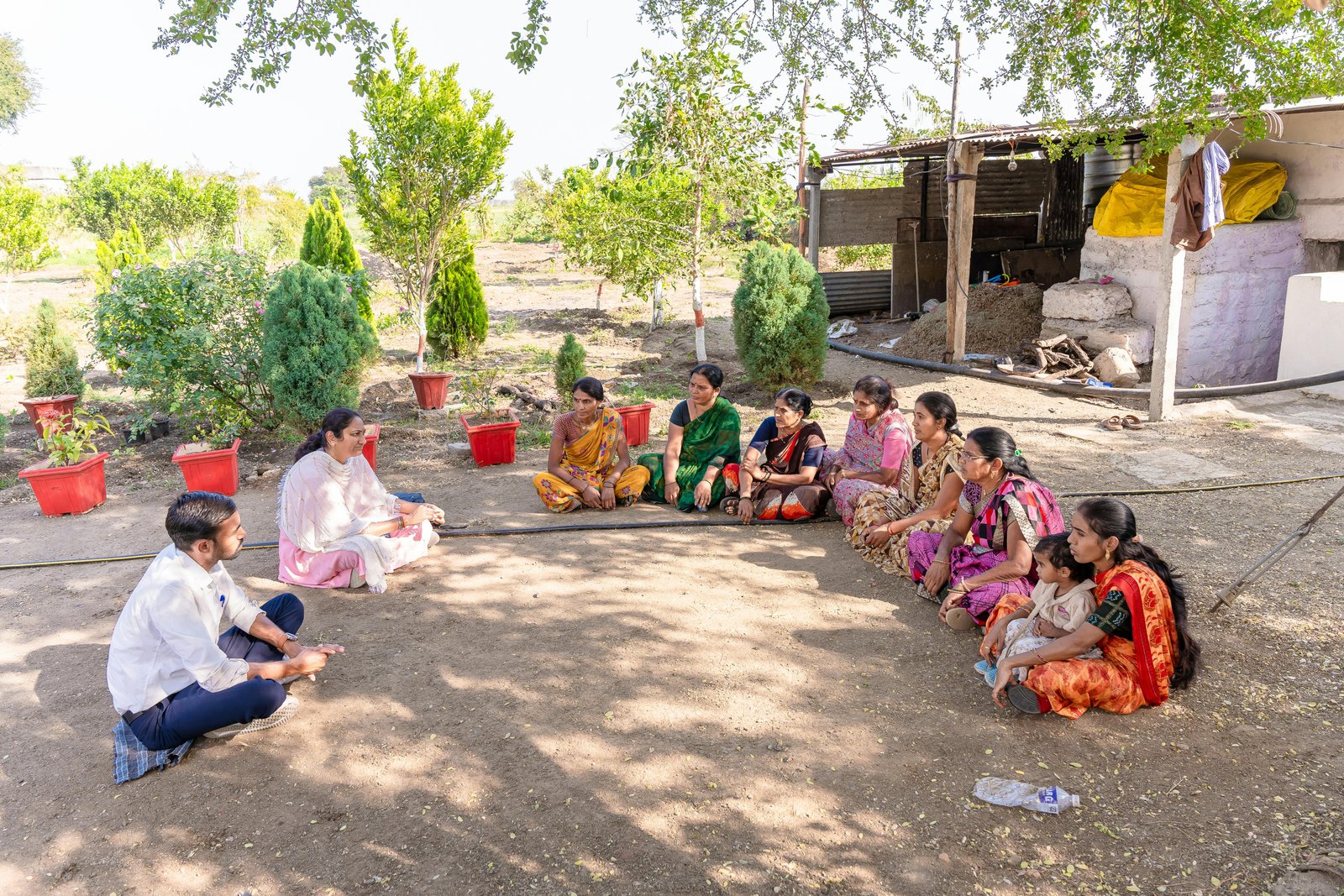 A group of women and children sit in a circle on the ground in a rural outdoor community space, appearing to participate in a community gathering or discussion session, with trees, vegetation, and a simple structure visible in the background.