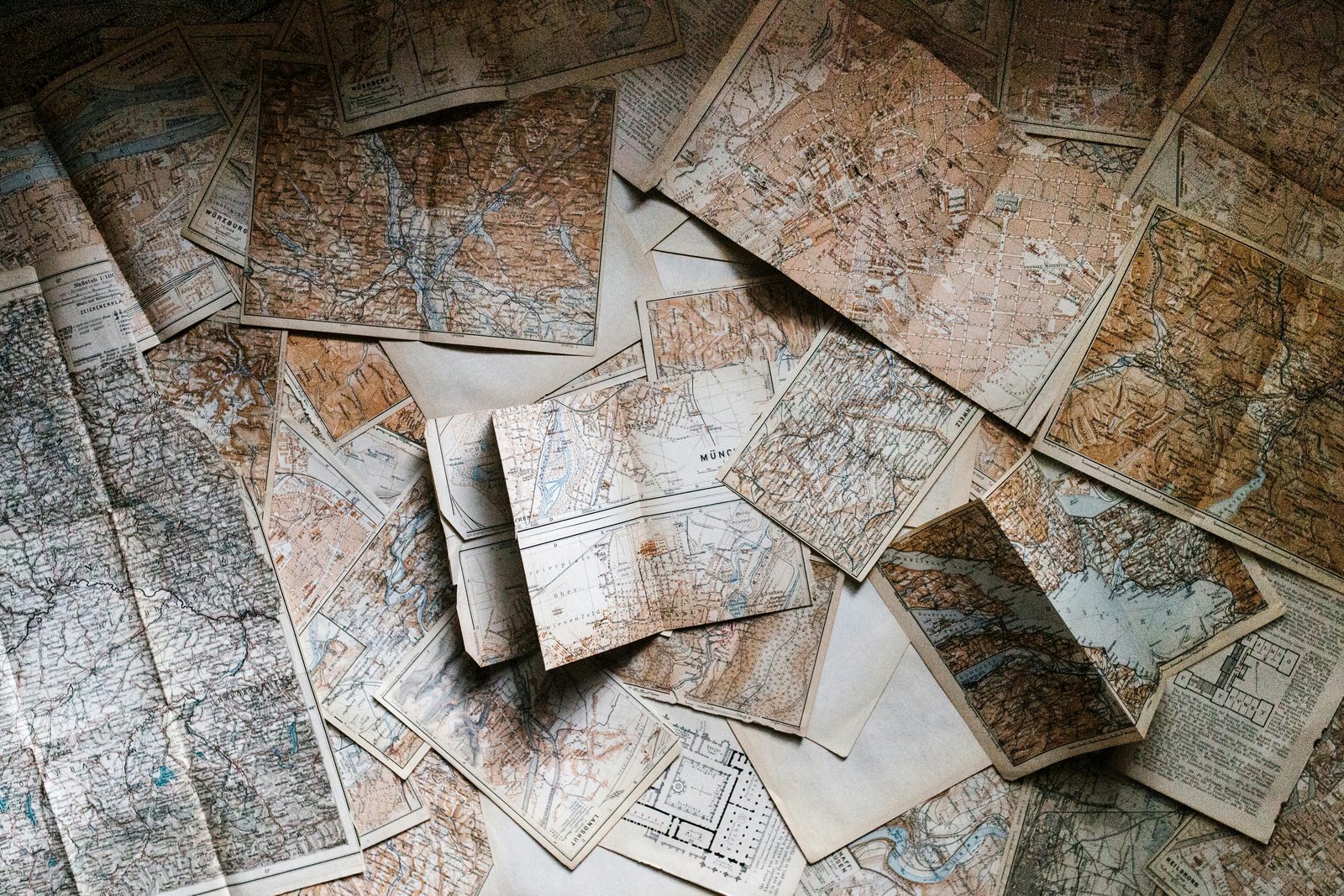 Overhead flat-lay photograph of numerous scattered vintage maps and historical documents with aged paper, some rolled, some stacked, displaying cartographic details and wear consistent with archival materials.