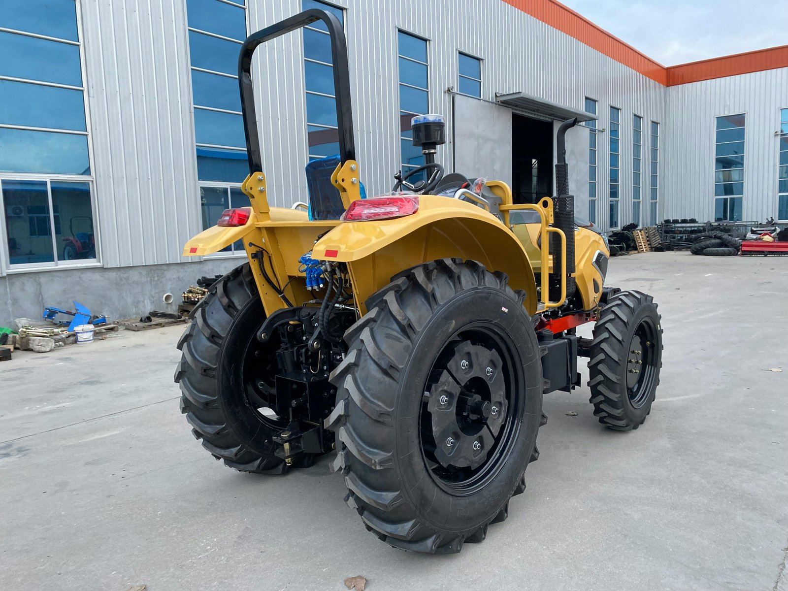 A yellow tractor with large black tires and a roll bar is parked in front of an industrial building with corrugated metal siding and blue-tinted windows.