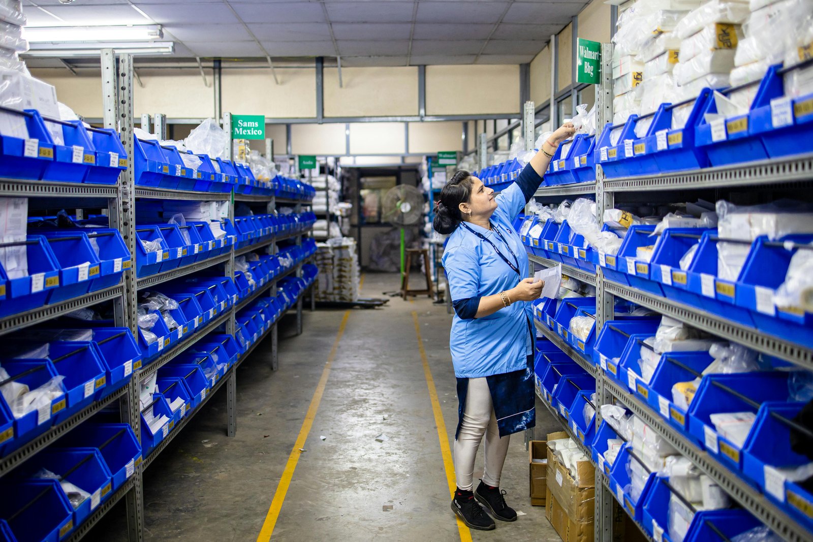 A worker in a light blue shirt stands in an industrial warehouse aisle between tall blue storage bins on metal shelving racks, reaching toward inventory items in a logistics distribution facility.