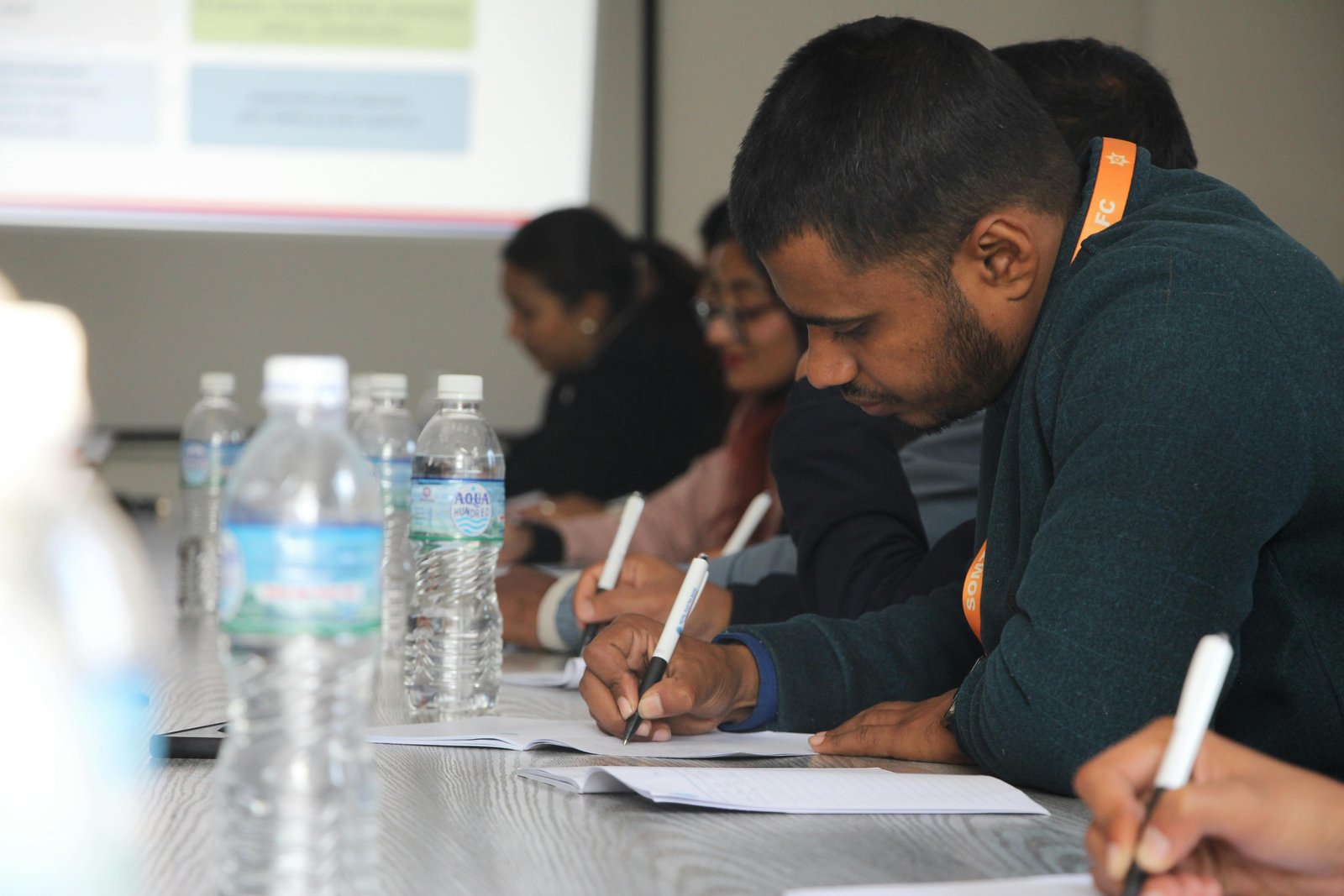 Multiple students seated at a table in a classroom setting, focused on writing notes during what appears to be a seminar discussion, with a projected presentation visible on a screen in the background and water bottles on the table.