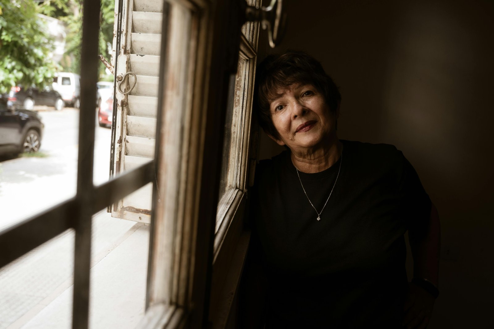 An adult person with dark hair wearing black clothing stands near a window in an interior space, gazing thoughtfully toward natural light coming from outside, with a calm and contemplative expression.