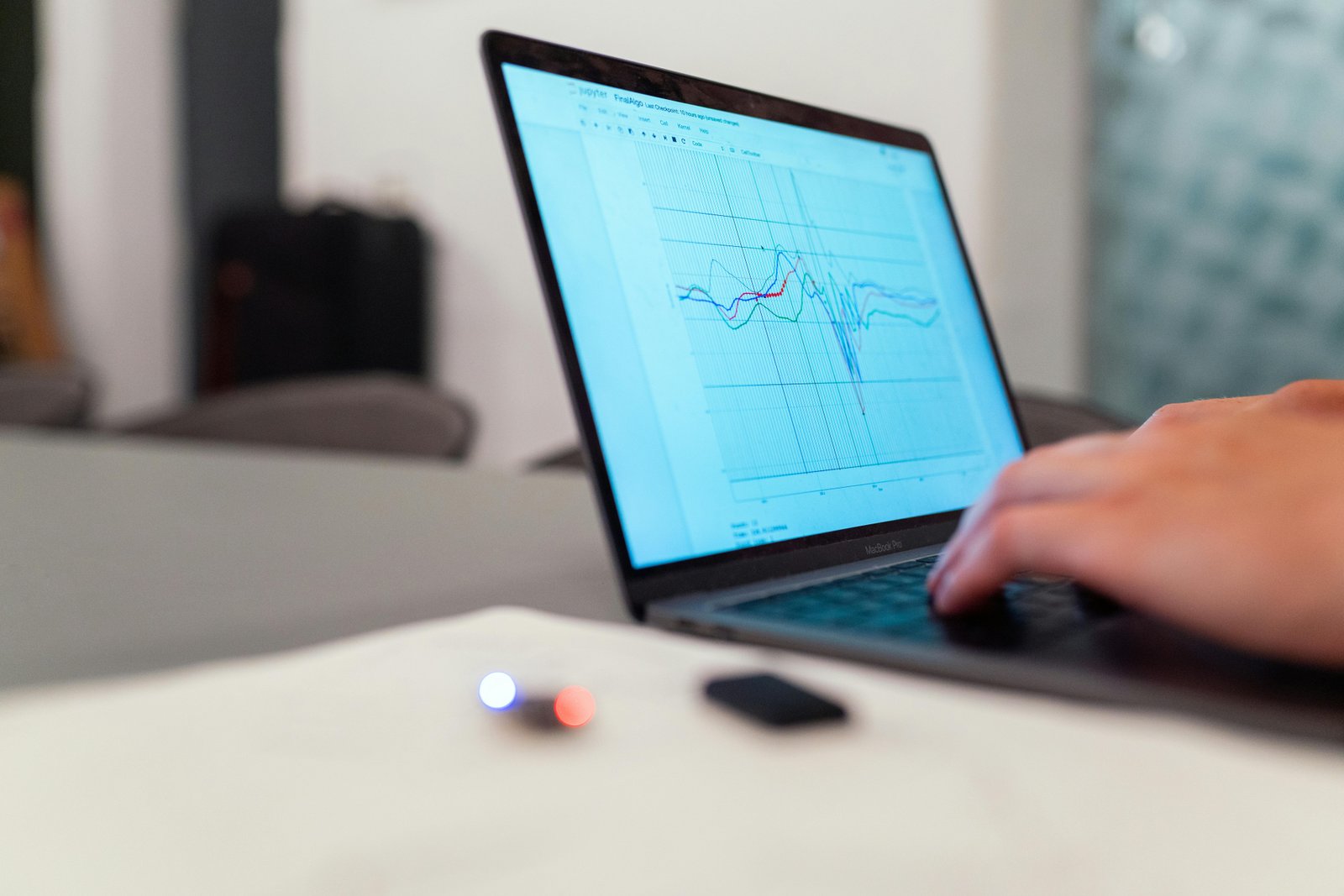 A person's hand operates a laptop displaying light blue graphs and network diagrams on its screen, positioned on a white desk in an office environment with blurred background elements.