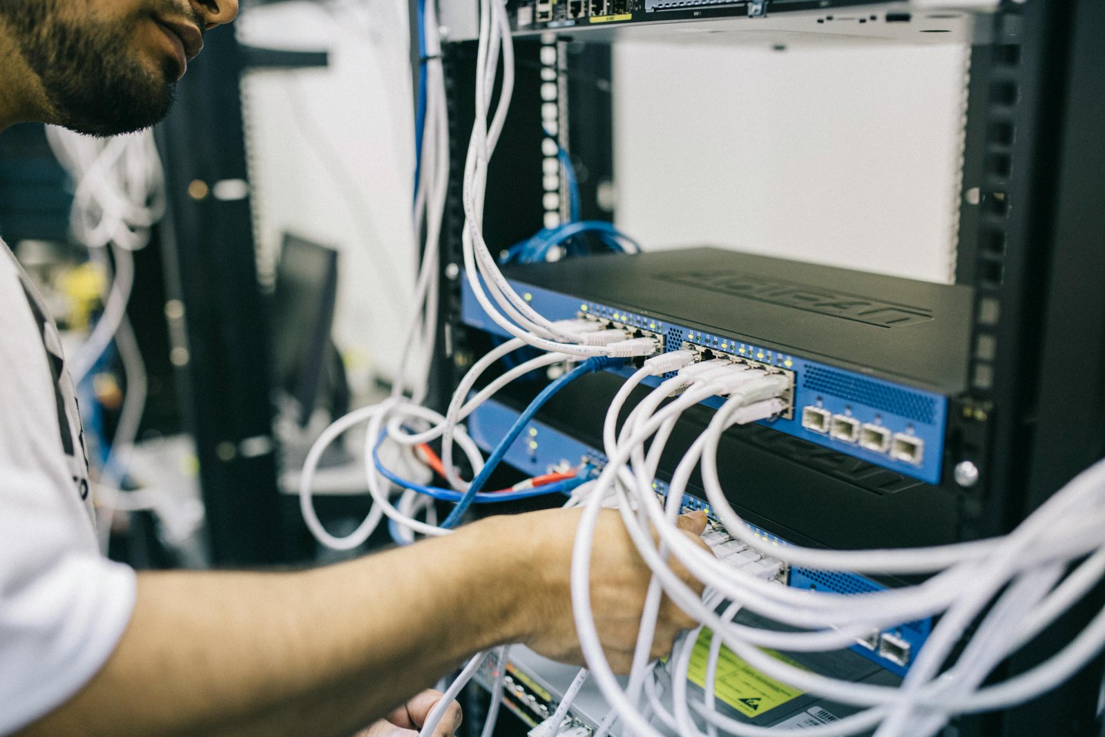 A technician's hands working with white network cables connected to blue and white networking equipment mounted in a server rack, with additional infrastructure visible in the background of what appears to be a data center facility.
