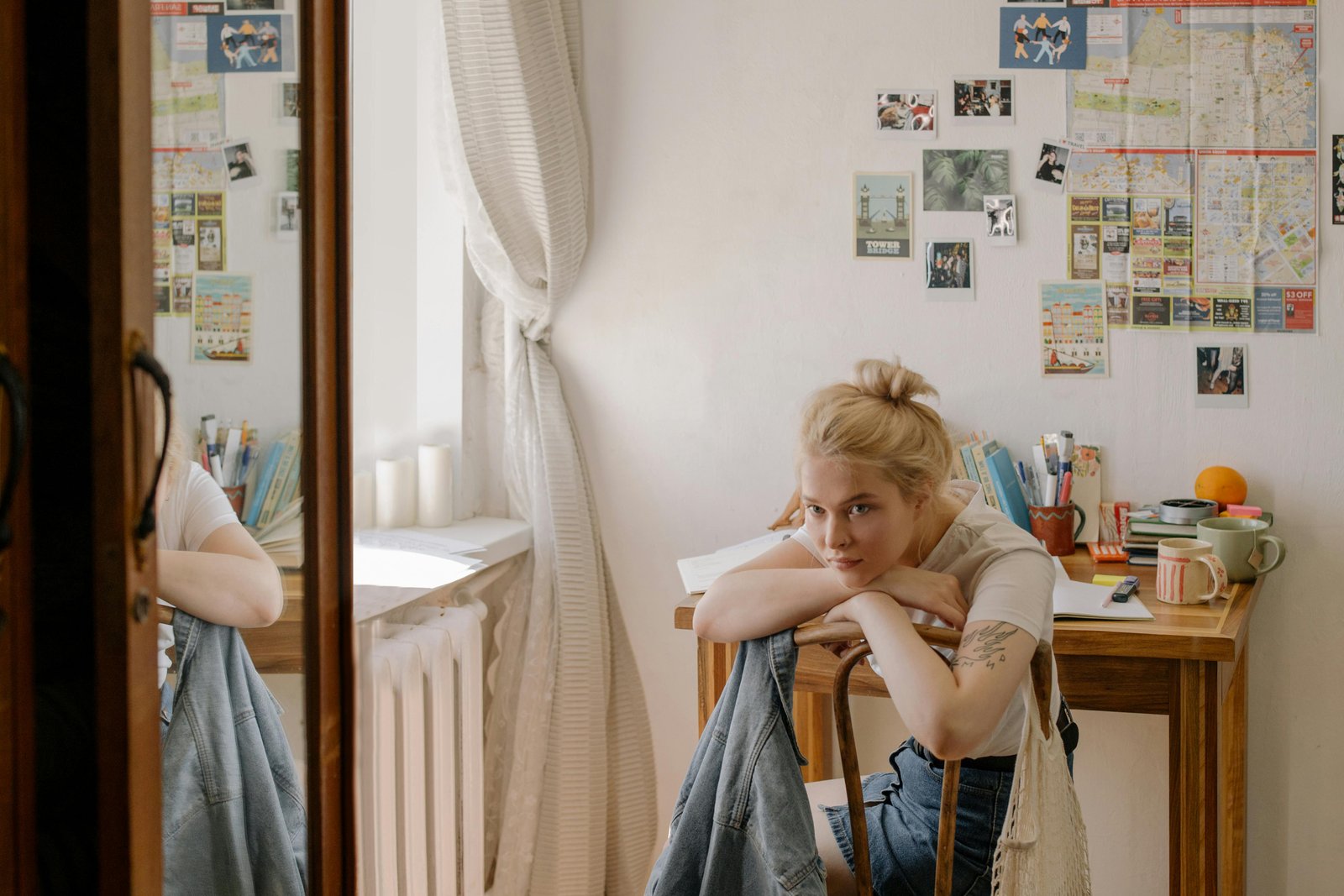 A young blonde woman sits on the floor leaning against a wooden desk in a modest room with white walls, wearing denim overalls, with photos and papers pinned to the wall behind her and a desk with various items visible.