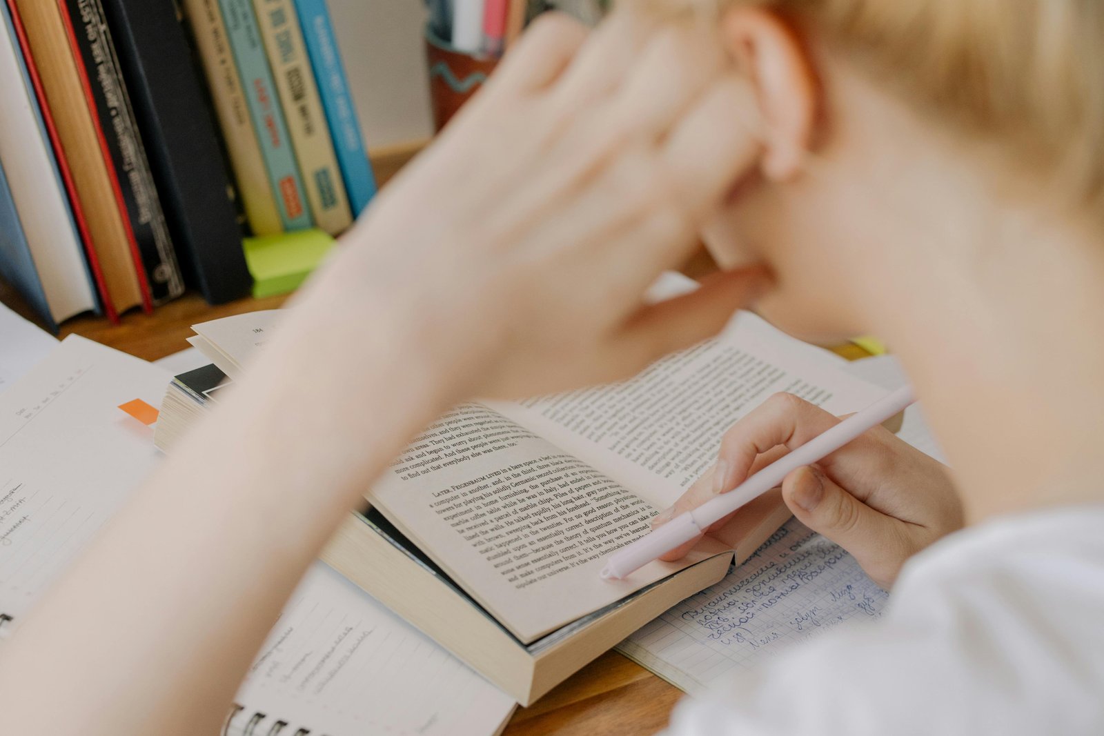 A person with red hair reading an open book at a desk covered with papers, with a bookshelf visible in the background.