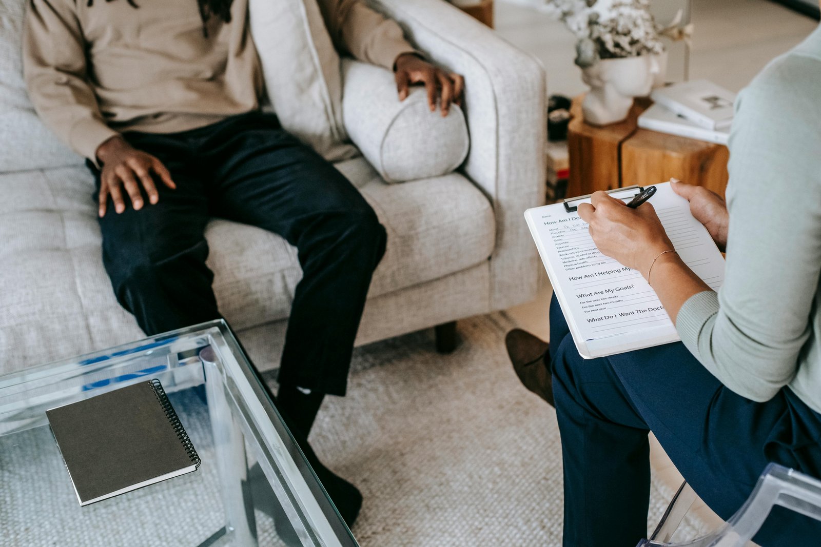 An overhead view of two people in a clinical consultation setting, with one person seated on a beige couch wearing dark pants and the other holding a clipboard with paperwork, with a glass table containing a tablet visible in the foreground.