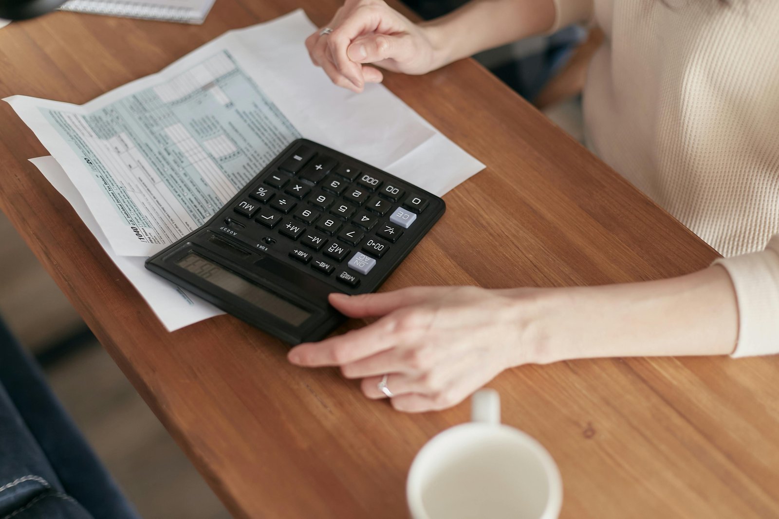 Two people sitting at a wooden table with a calculator and papers, with hands positioned near the calculator and a white mug visible in the foreground.