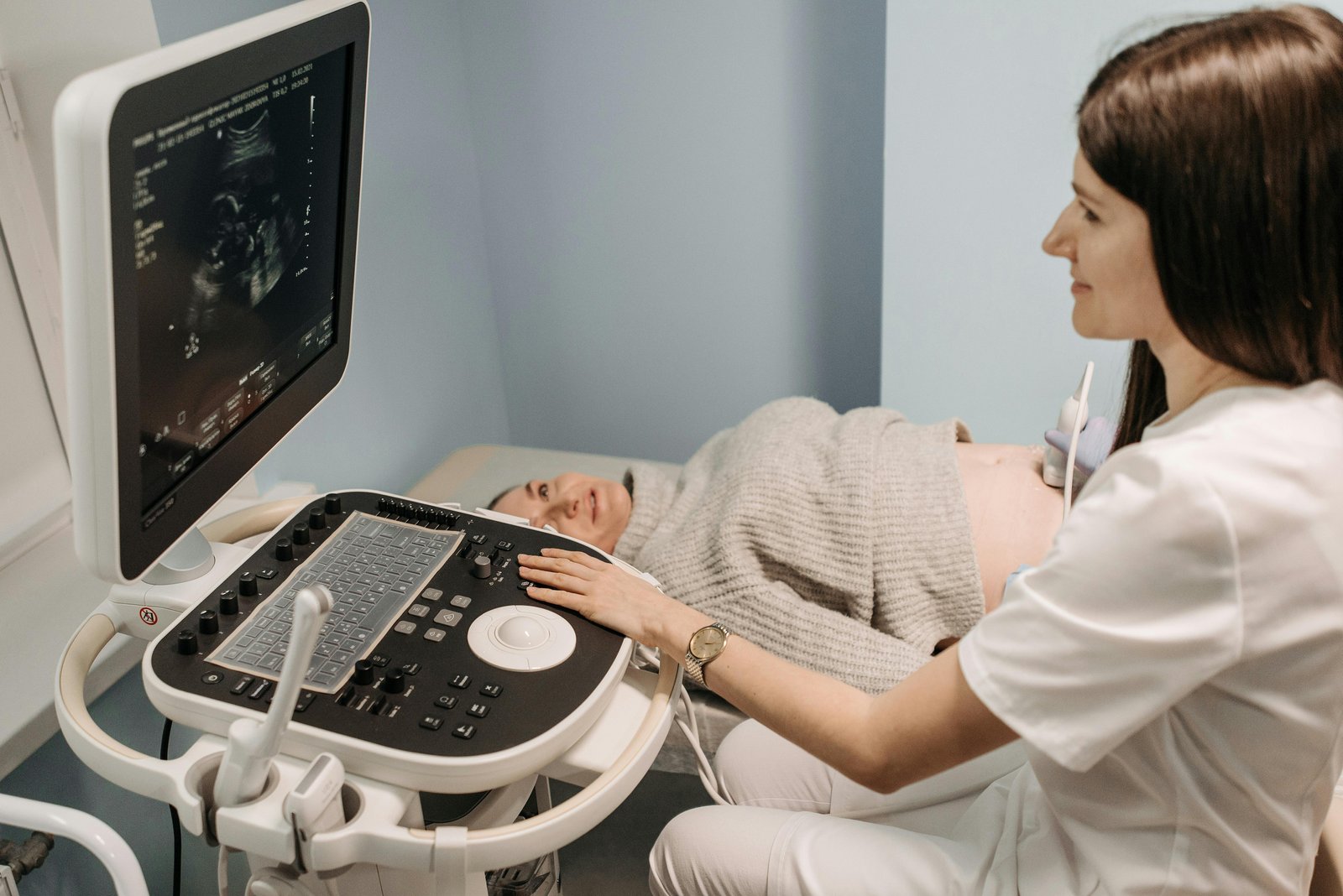 A medical professional in white attire performs an ultrasound examination on a pregnant woman who is lying back while the professional operates the ultrasound machine's control panel, with the ultrasound monitor displaying diagnostic images in the background.