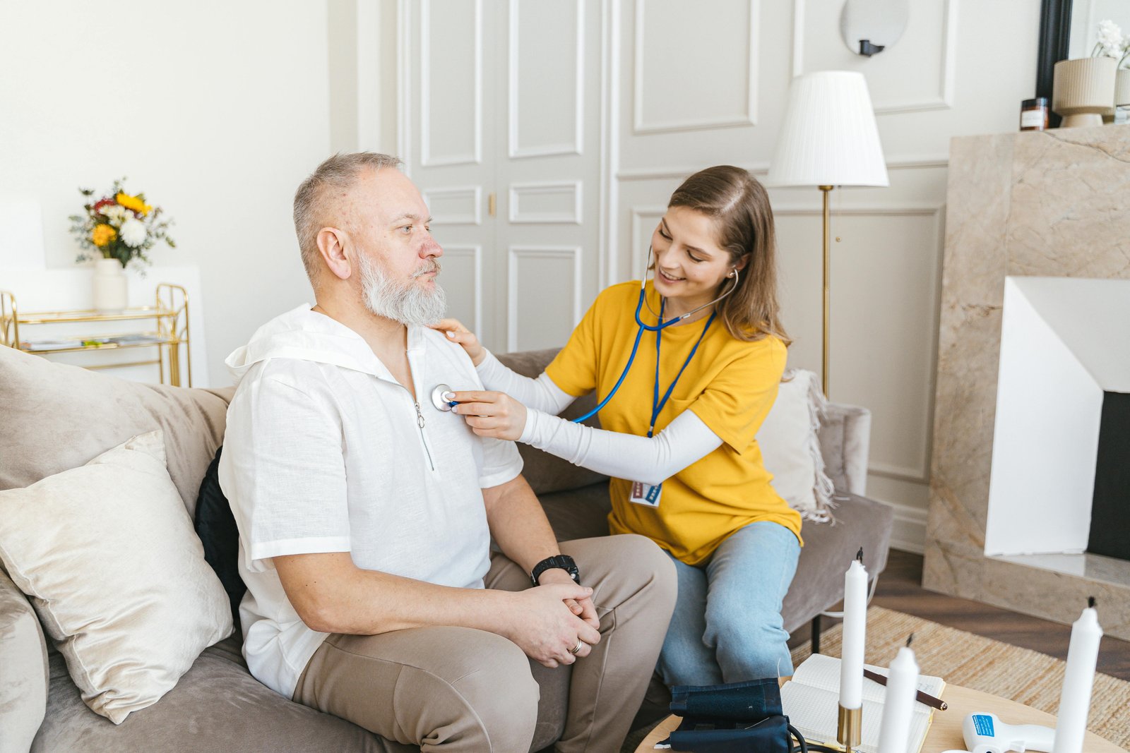A healthcare worker in a yellow top uses a stethoscope to examine an older man with a white beard who is seated on a beige couch in a living room.