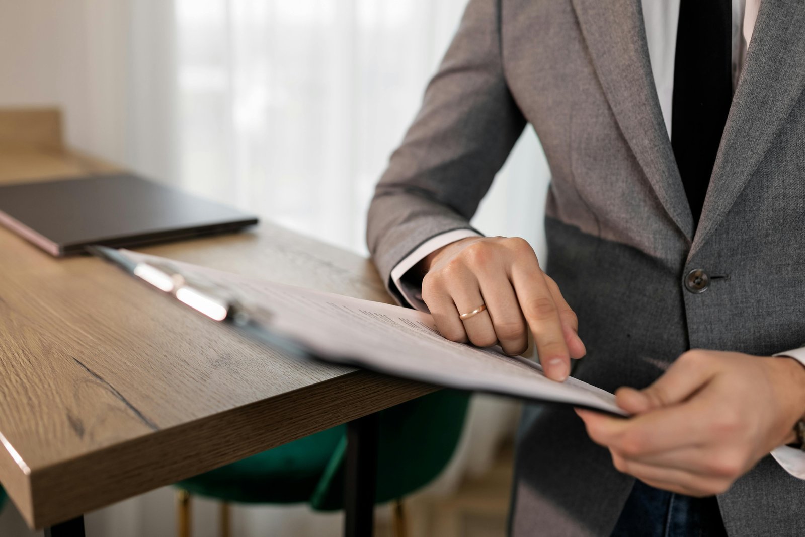 A person in a gray business blazer sits at a wooden desk reviewing documents with a pen in hand, with a laptop and window visible in the background.