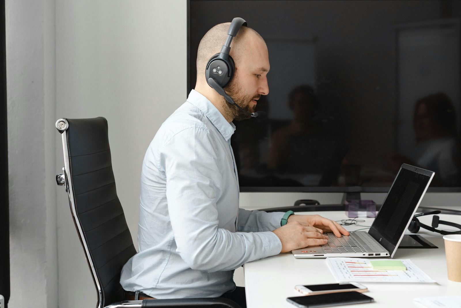 A man wearing a headset sits at a desk working on a laptop in an office environment.