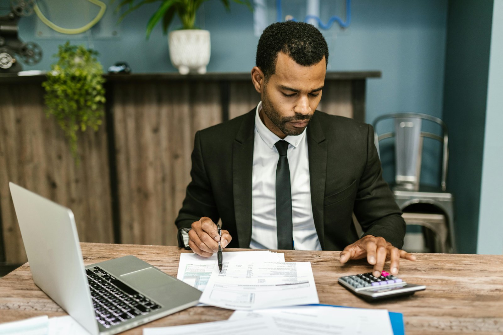 A man in a dark business suit and tie sits at a wooden desk in a modern office with blue-green walls, writing on financial documents while working at a laptop, with a calculator and other office supplies visible on the desk.