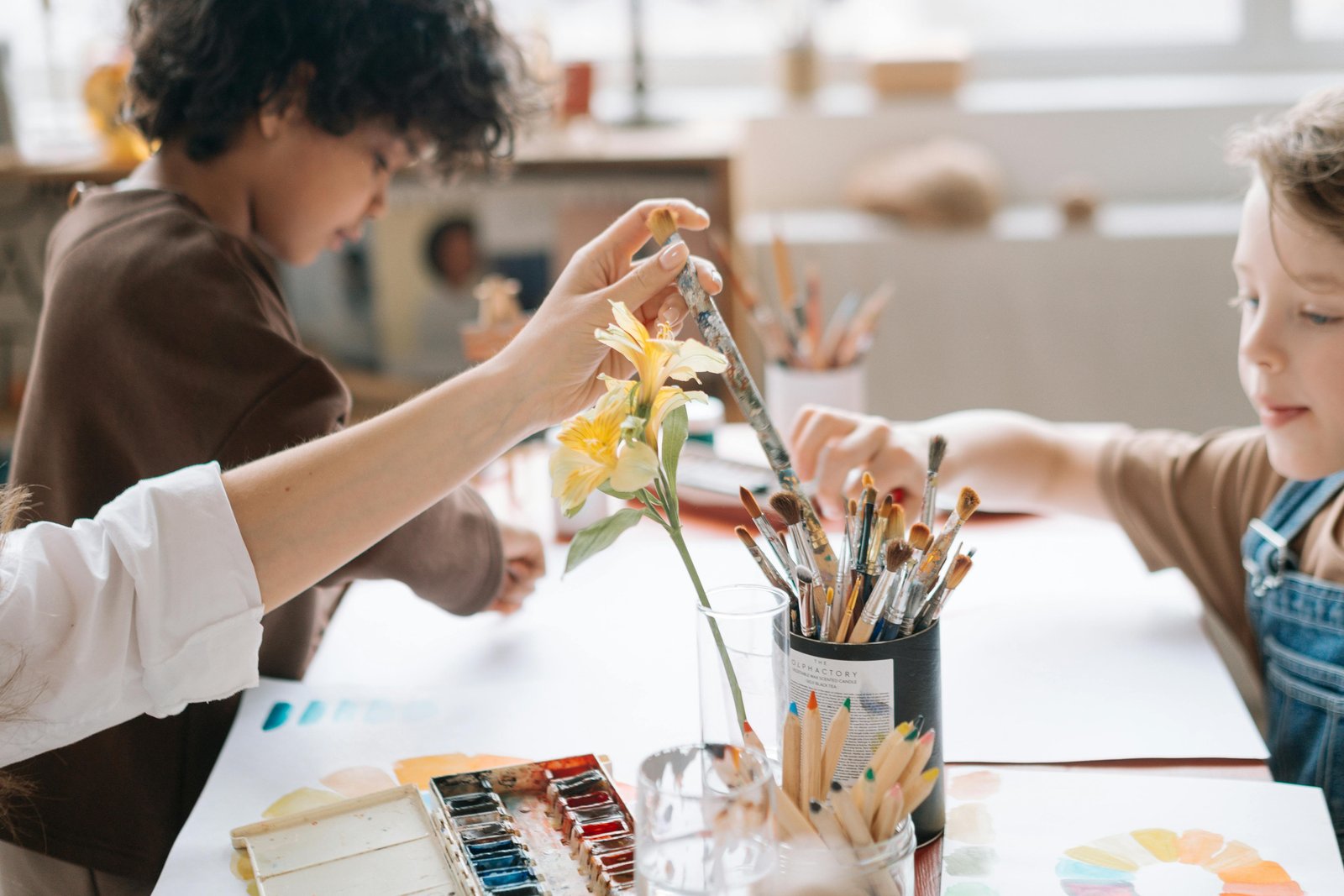 Children in a bright classroom setting are engaged in collaborative art-making at a table, with one child holding up a yellow flower artwork while others work nearby with paintbrushes, paint supplies, and creative materials visible on the table.