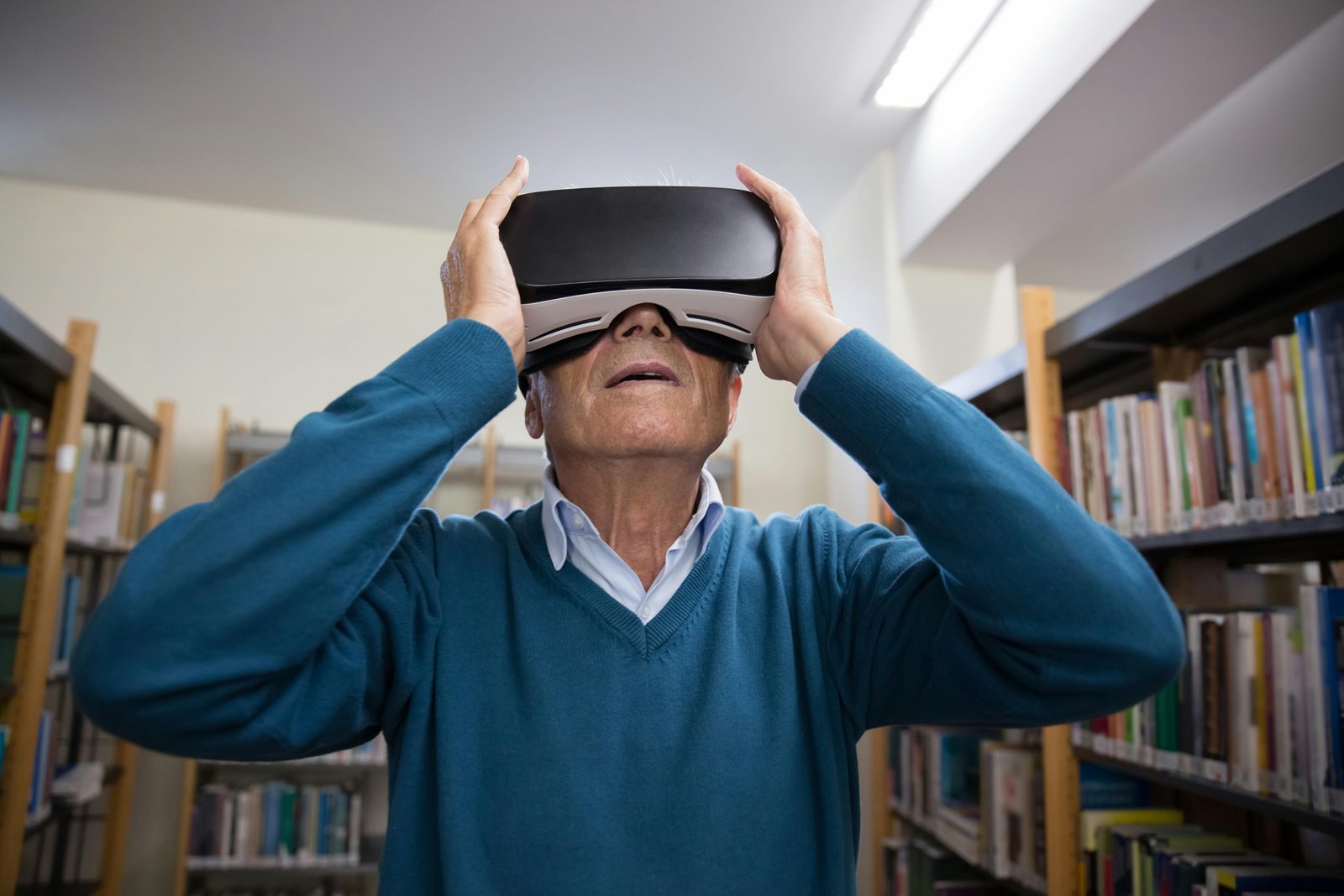 An older adult man wearing a blue sweater stands in a library with bookshelves visible in the background, holding a black VR headset up to his face with both hands.