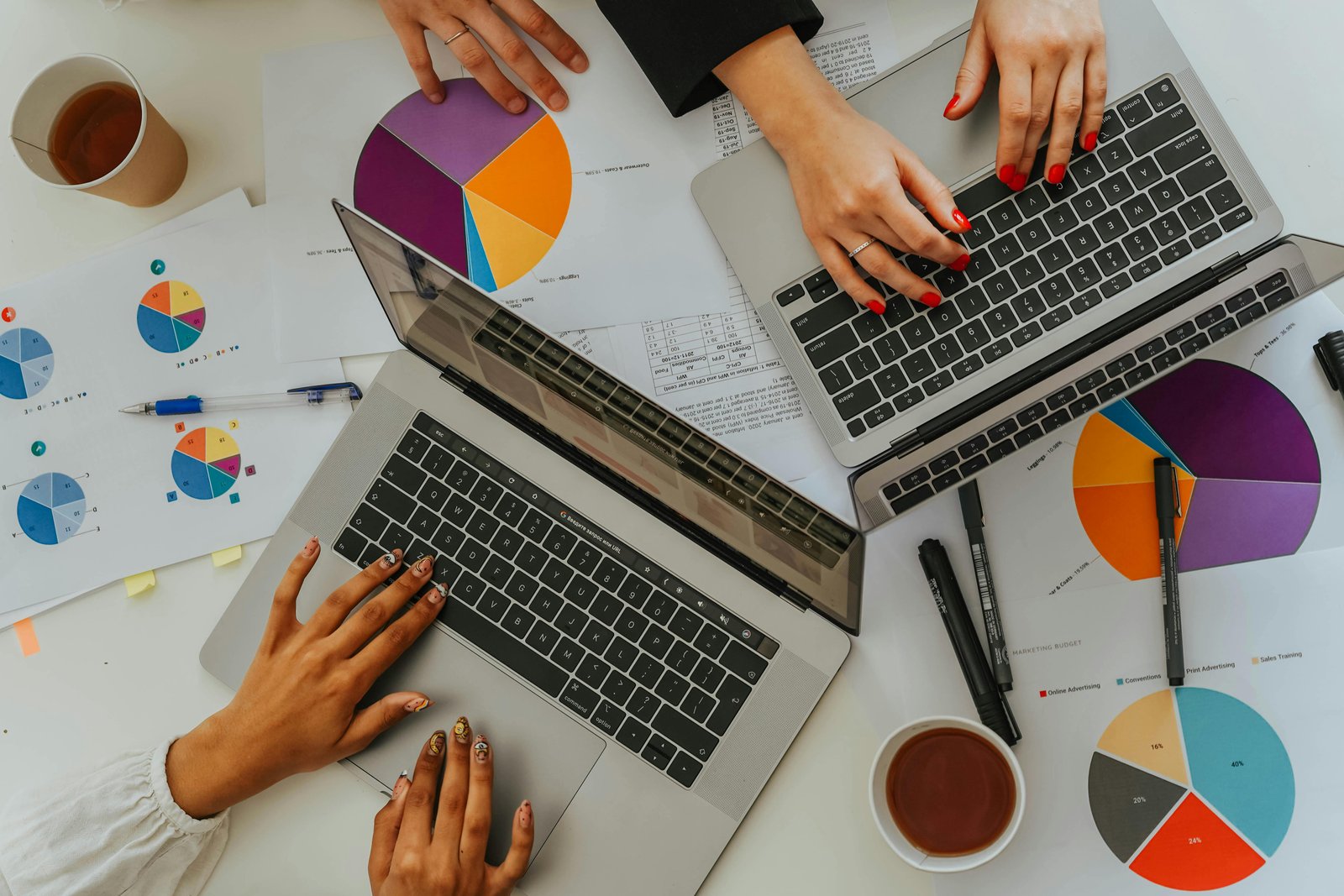 An overhead view of two people working on laptops at a white desk covered with colorful pie charts, printed documents, pens, and cups of coffee.