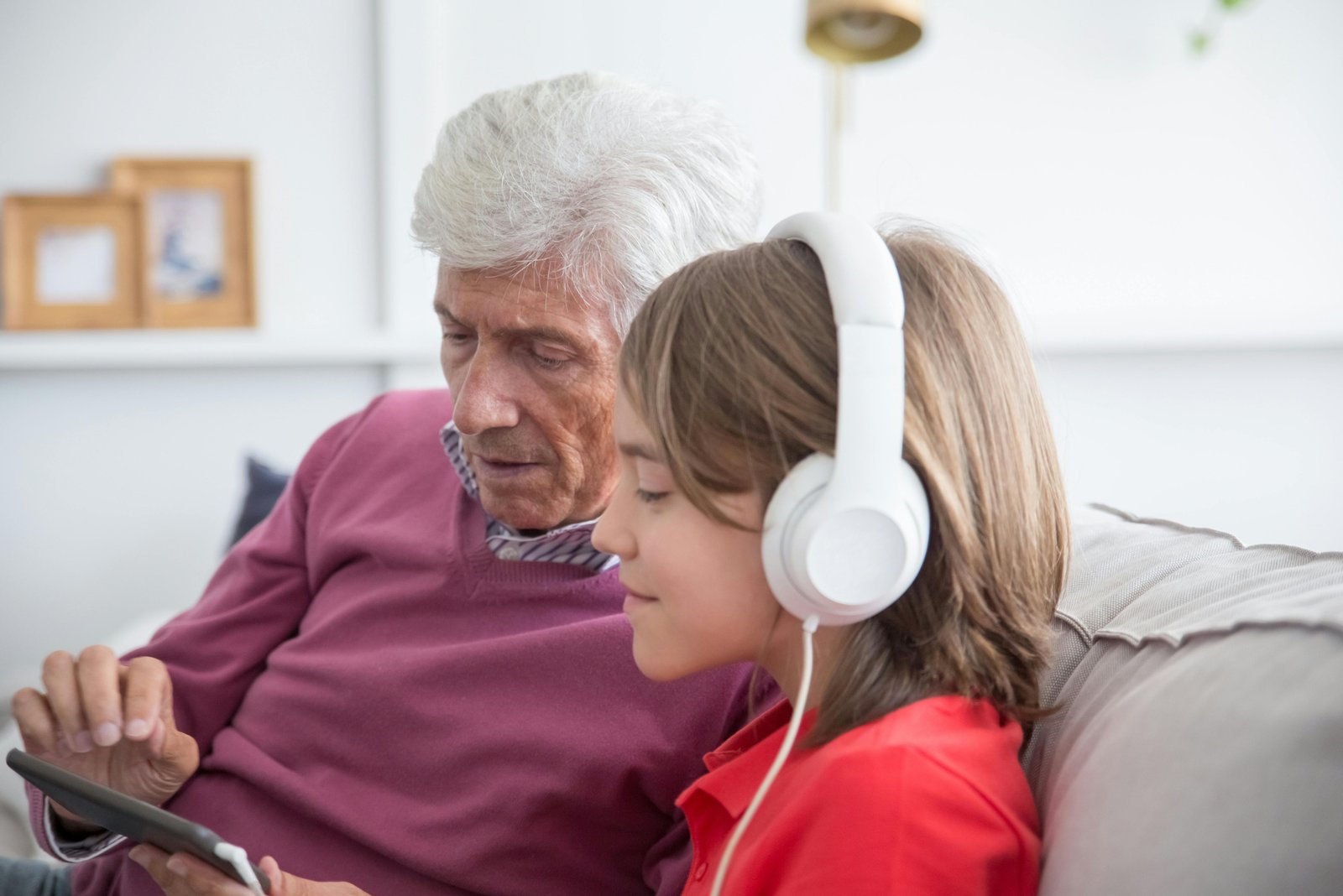 An older man in a burgundy sweater and a young girl wearing white headphones sit together on a couch, looking at a tablet device in a bright, modern living room setting.