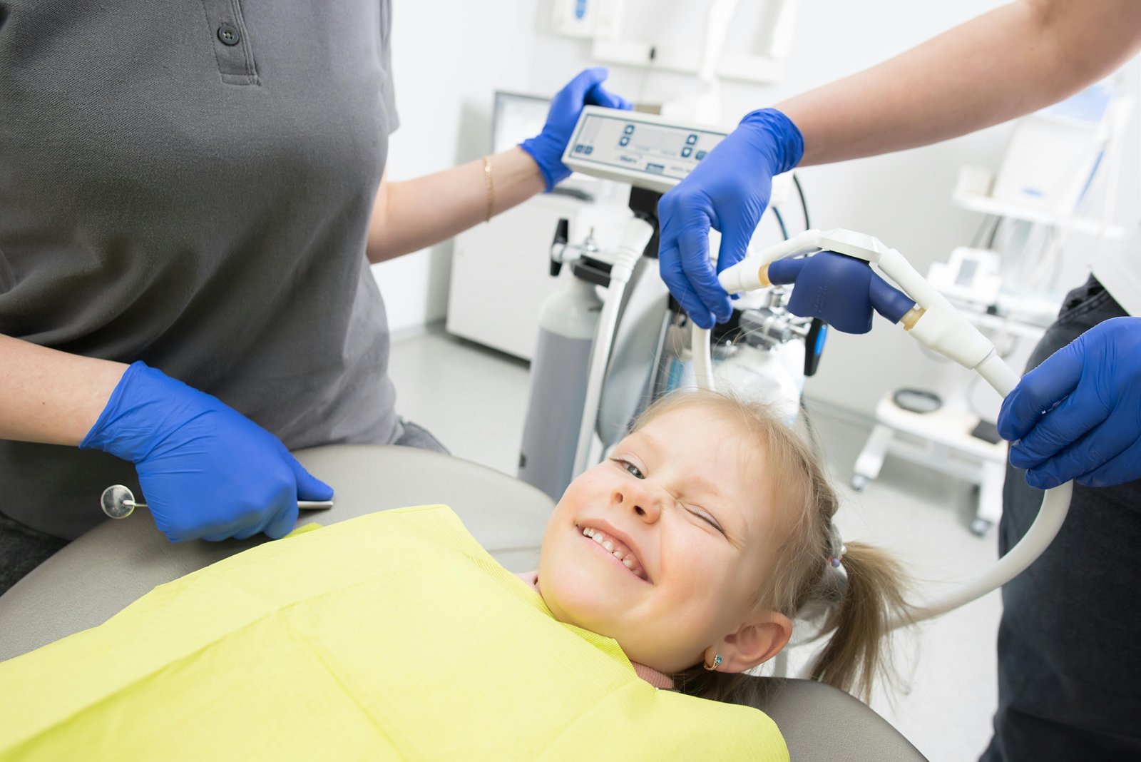 A young child in a yellow dental bib sits in a dental chair while a healthcare provider wearing blue gloves and gray clothing performs an intraoral dental examination with dental instruments, with dental equipment visible in the background.