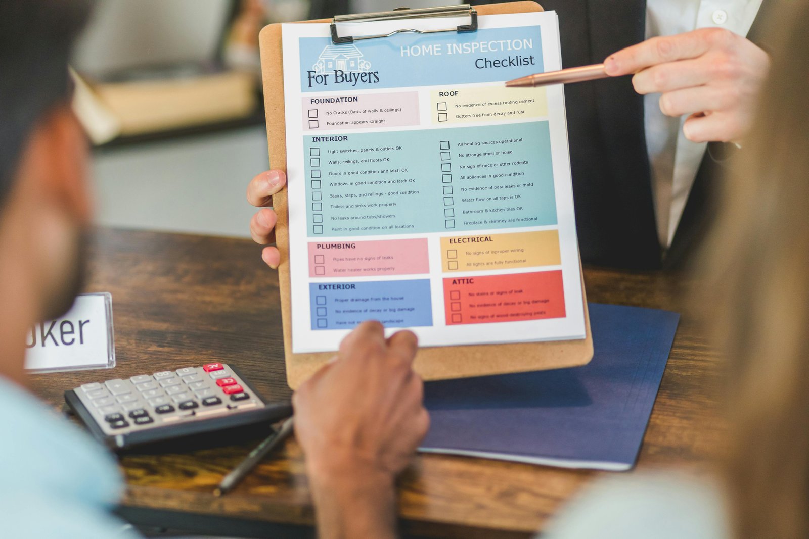 A person holds a clipboard with a home inspection checklist while another person points at it with a pen, with a calculator visible on the table.