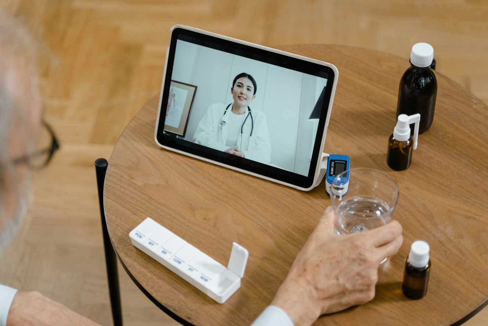 A person's hands holding a glass of water at a wooden desk during a video call with a healthcare professional displayed on a tablet screen, with medical documents and medication bottles visible in the foreground.