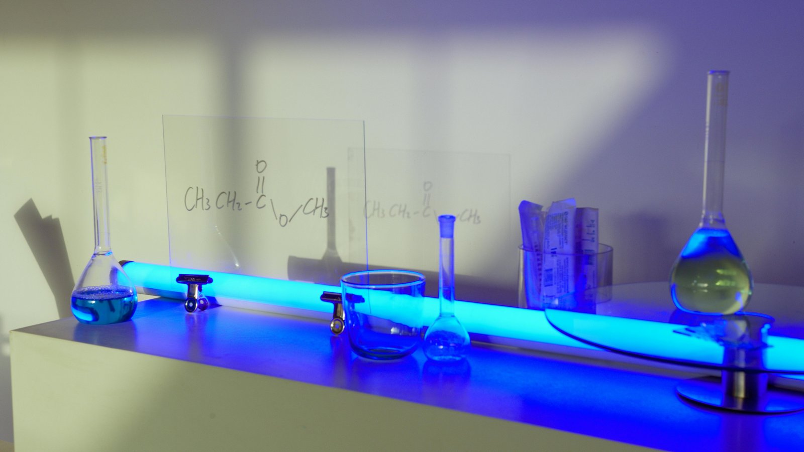 A laboratory setup with glass beakers, test tubes, and an Erlenmeyer flask containing blue liquid, illuminated by blue light on a shelf with chemical formulas written on cards in the background.