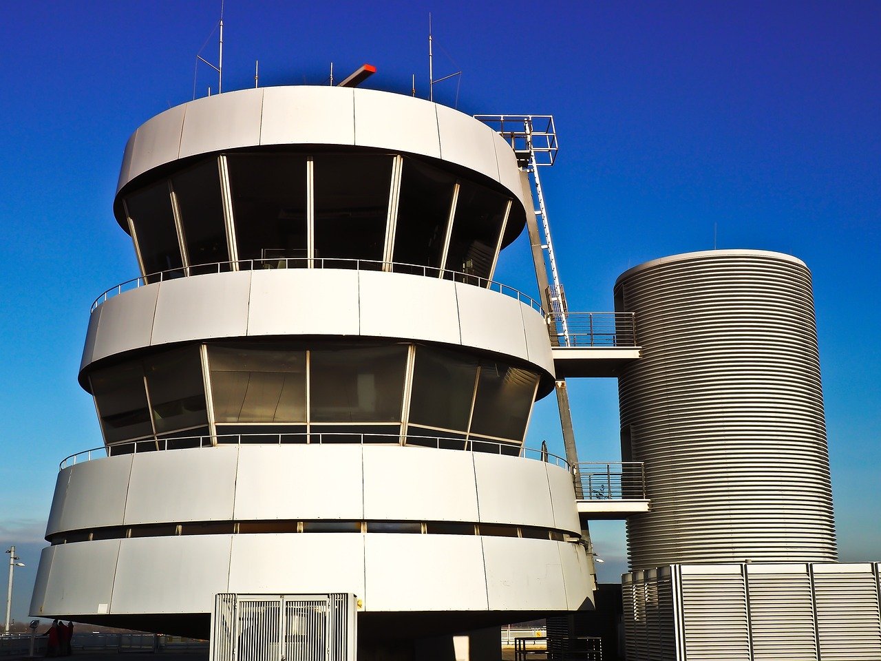 A modern airport control tower with distinctive cylindrical architecture featuring a multi-level observation deck and attached cylindrical structure against a clear blue sky.