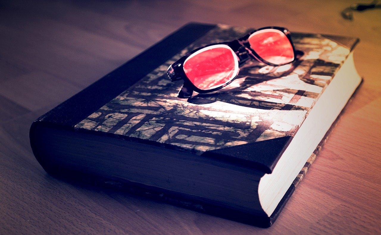 An overhead view of a desk with printed documents, a pair of pink-framed eyeglasses resting on top of the papers, and what appears to be a dark blue book or folder, arranged on a wooden surface in natural lighting.