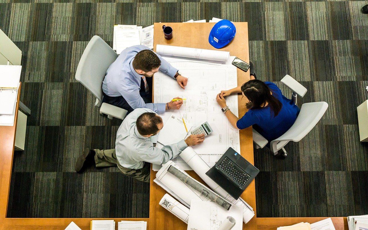 An overhead view of three people seated around a wooden table covered with architectural plans, blueprints, a laptop, a blue hard hat, and office supplies on a striped carpet floor.