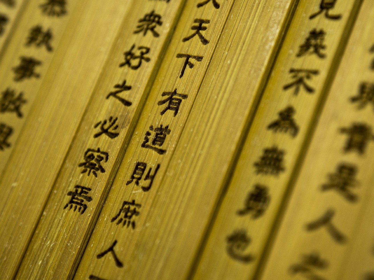 Close-up angled view of multiple wooden sticks or strips with traditional Chinese characters printed in black ink on a natural tan/beige wooden surface, arranged in parallel rows.