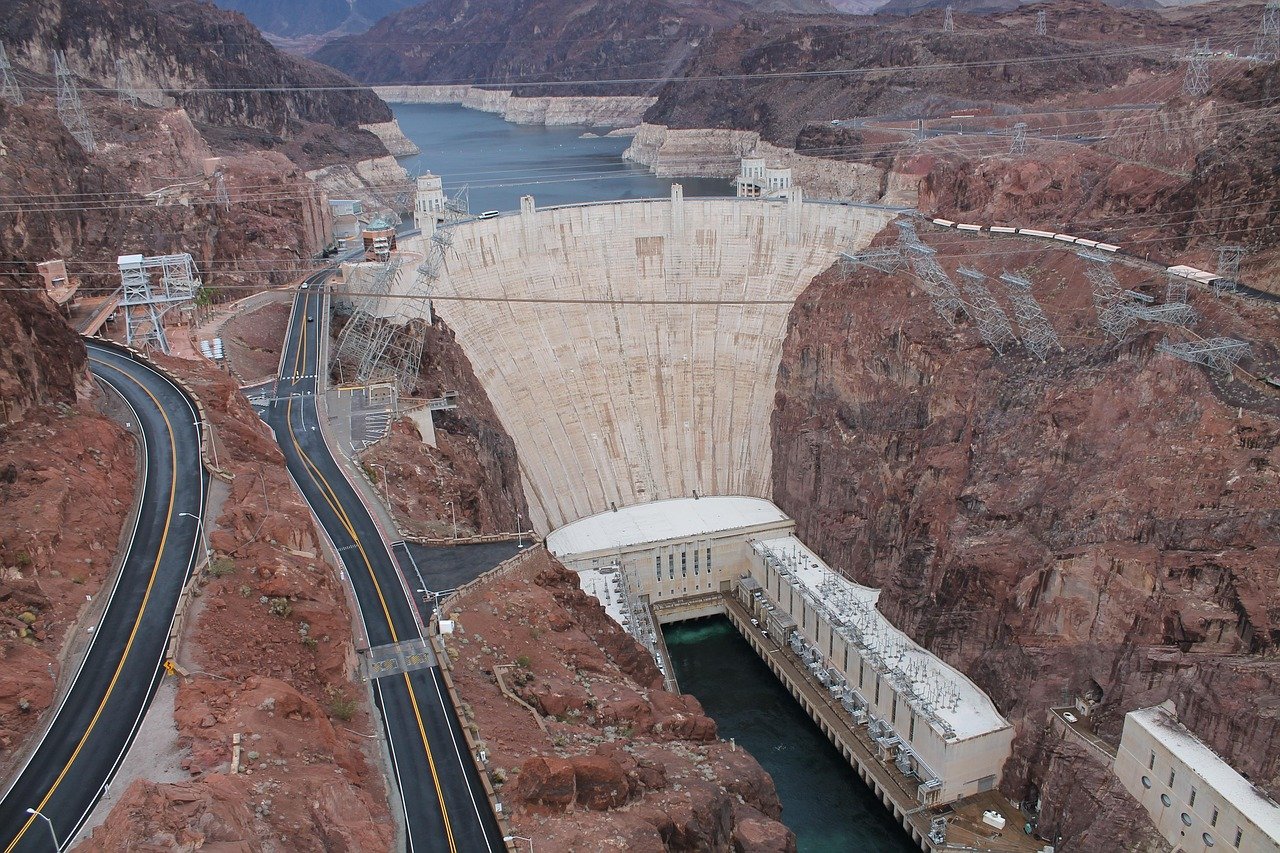 Aerial photograph of a large concrete dam spanning a river canyon with arid, rocky terrain, showing the reservoir behind the dam, surrounding irrigation infrastructure, and topographic landscape features.