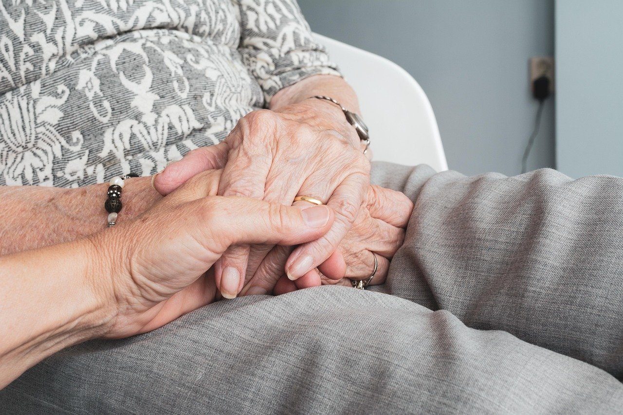 An older adult's hands gently hold and support a young child's hand in what appears to be a domestic interior setting, suggesting an intergenerational caregiving moment.