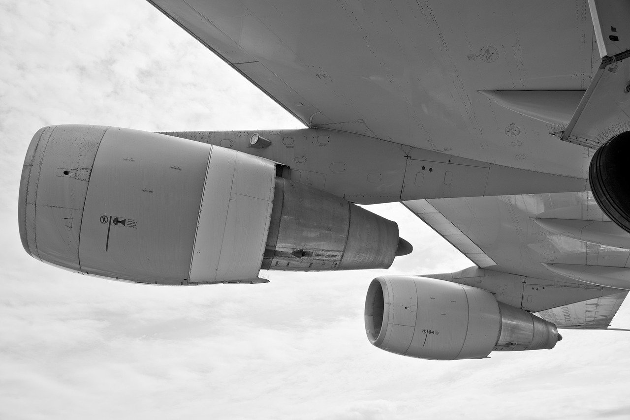Black and white photograph showing an aircraft wing and two jet engines viewed from below against a cloudy sky background with stratiform cloud layers visible.