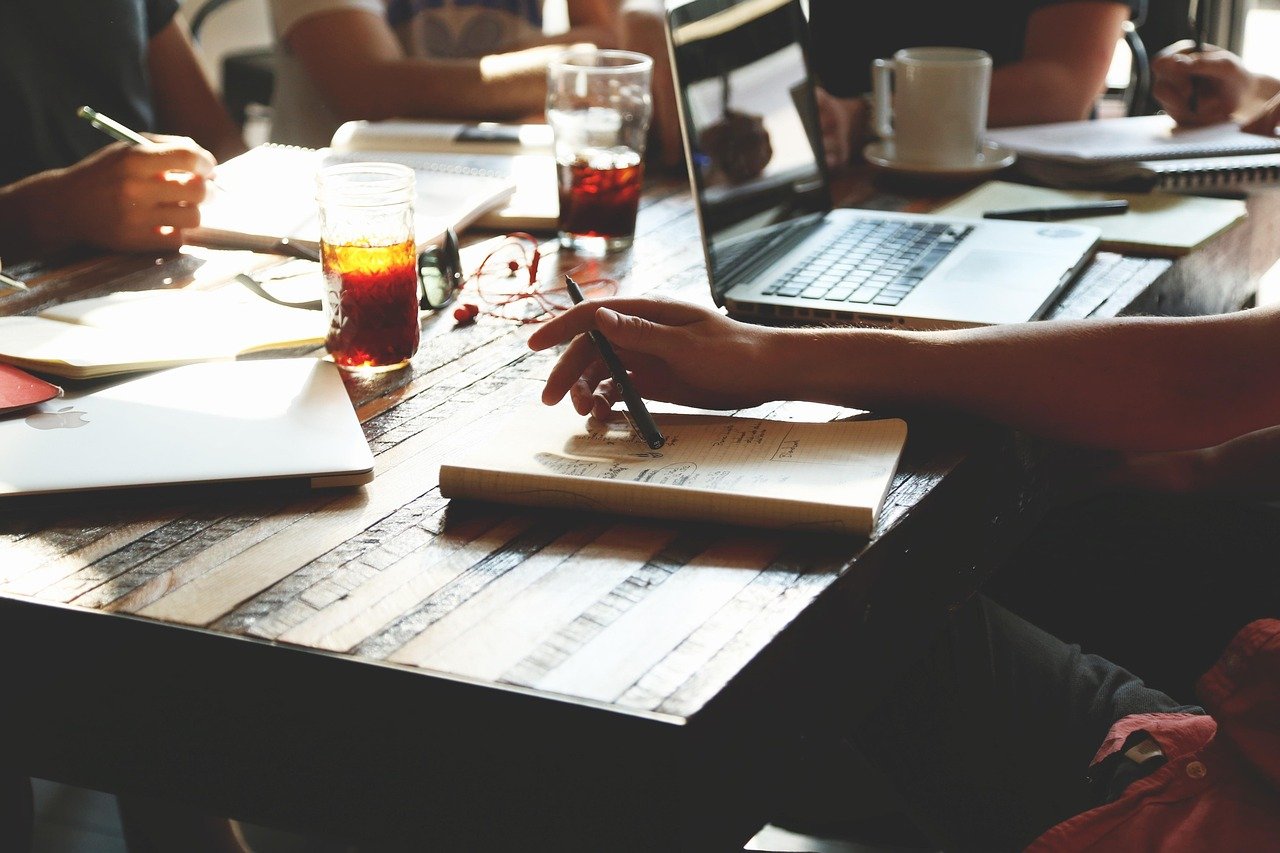 A overhead view of a conference table with scattered papers, documents, writing implements, and office materials spread across a dark surface, with people seated around the table in what appears to be a professional meeting or research session.
