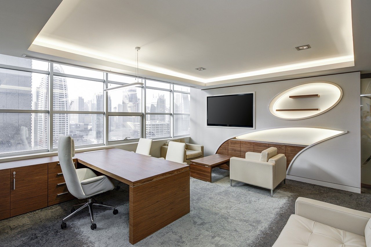 A modern government office interior featuring a wooden desk with a computer monitor, ergonomic office chair, floor-to-ceiling windows with city views, contemporary ceiling design with curved lighting elements, and professional institutional architecture in a landscape orientation.