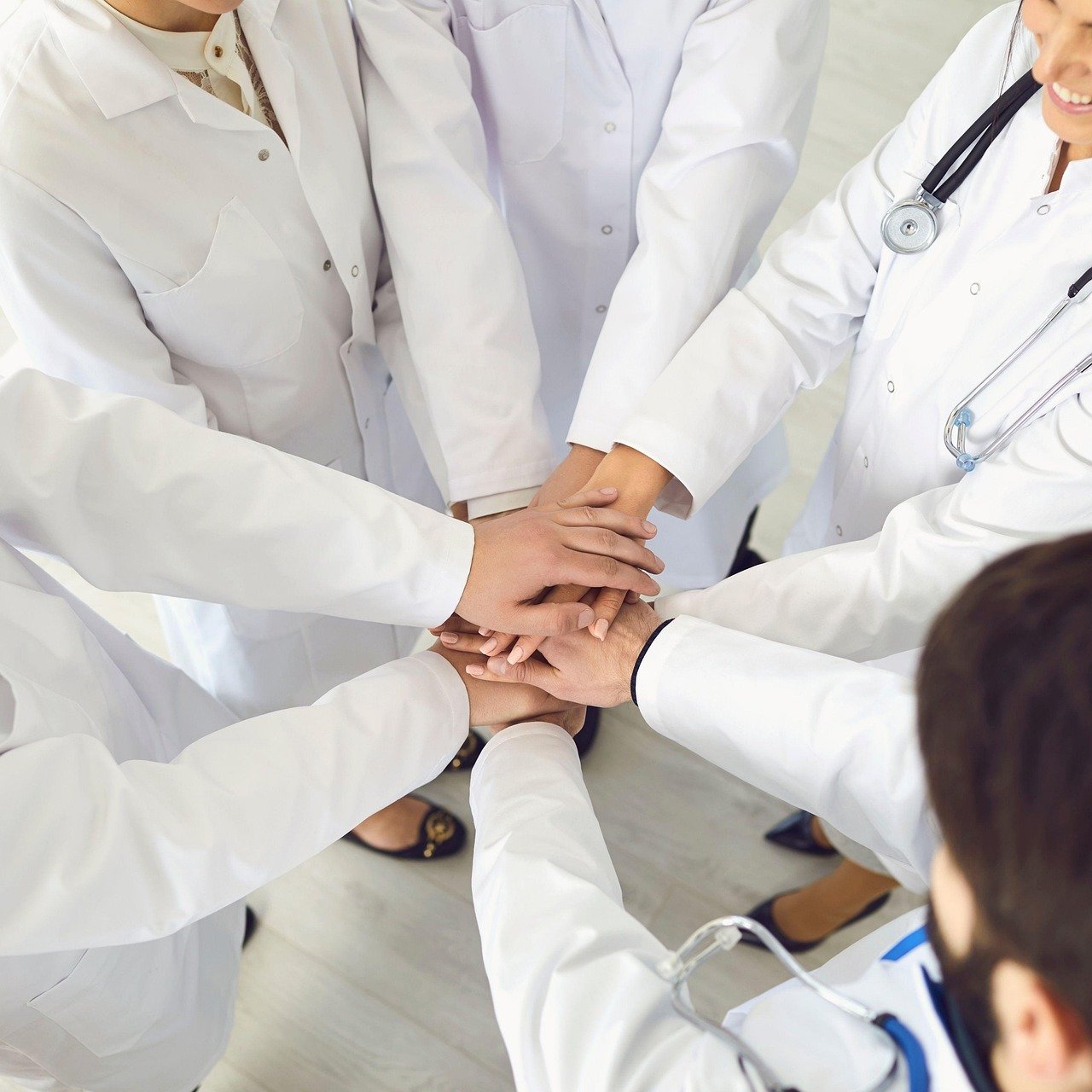 A group of healthcare professionals in white coats stand in a circle with their hands joined together at the center, viewed from below in an overhead composition.