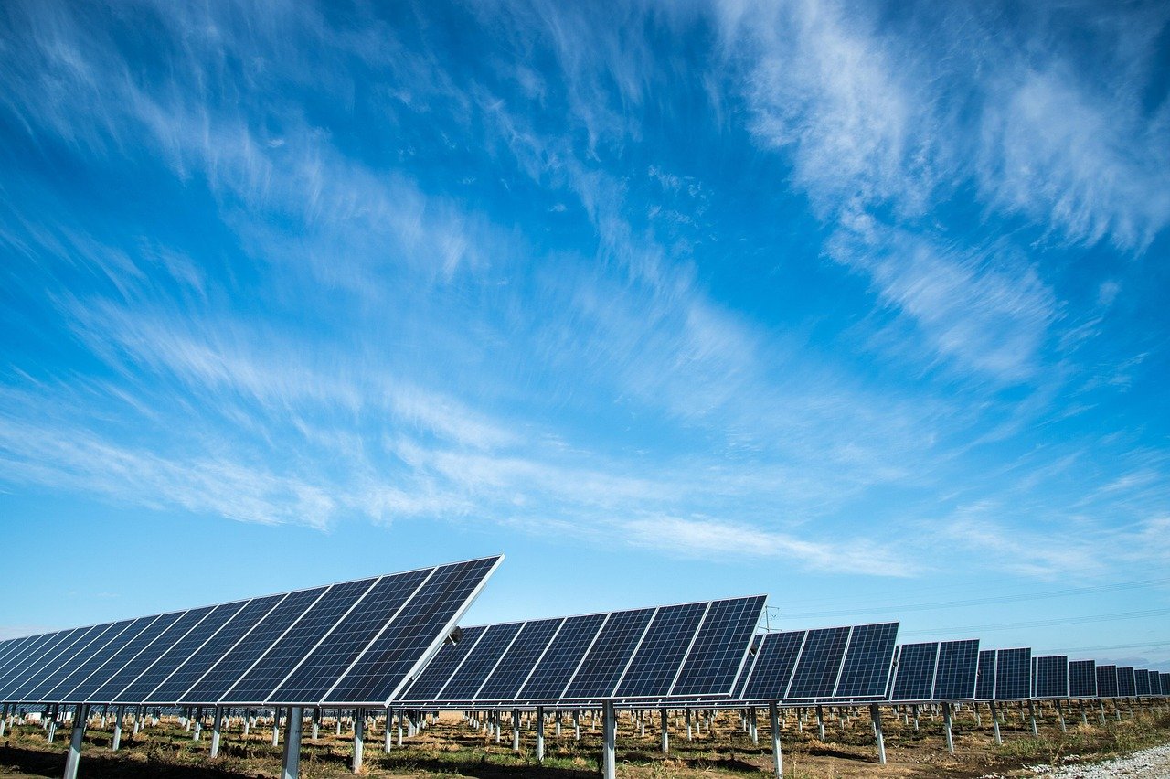 A wide landscape view of a large solar panel array stretching across the ground beneath a bright blue sky with wispy white clouds.
