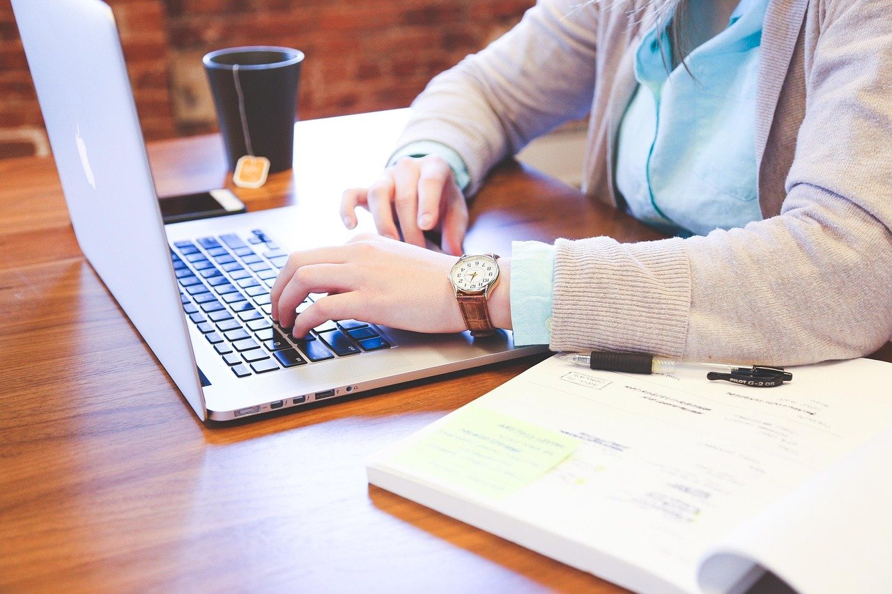 A person wearing light blue clothing works at a laptop on a wooden desk while reviewing documents, with a mobile device and pen visible in a modest workspace.