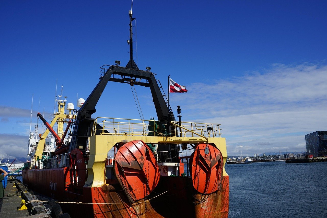 A large industrial vessel with orange and red coloring and multiple cranes or lifting equipment sits on calm blue-green ocean water under a clear sky, with a distant coastline visible on the horizon.