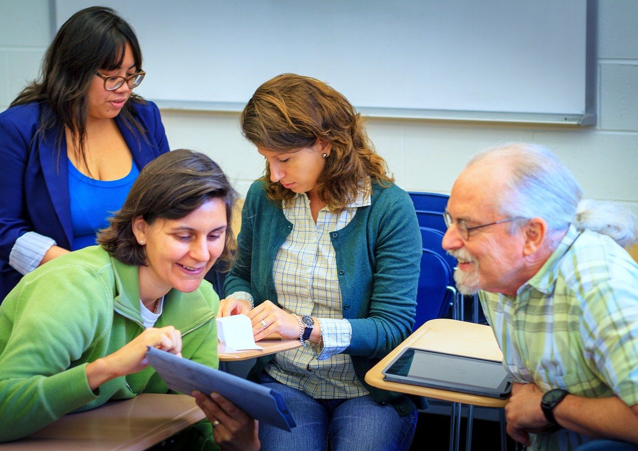 Four young students of mixed ages gathered around a table in a classroom, collaboratively engaged with educational materials and cards, with a periodic table chart visible on the classroom wall in the background.