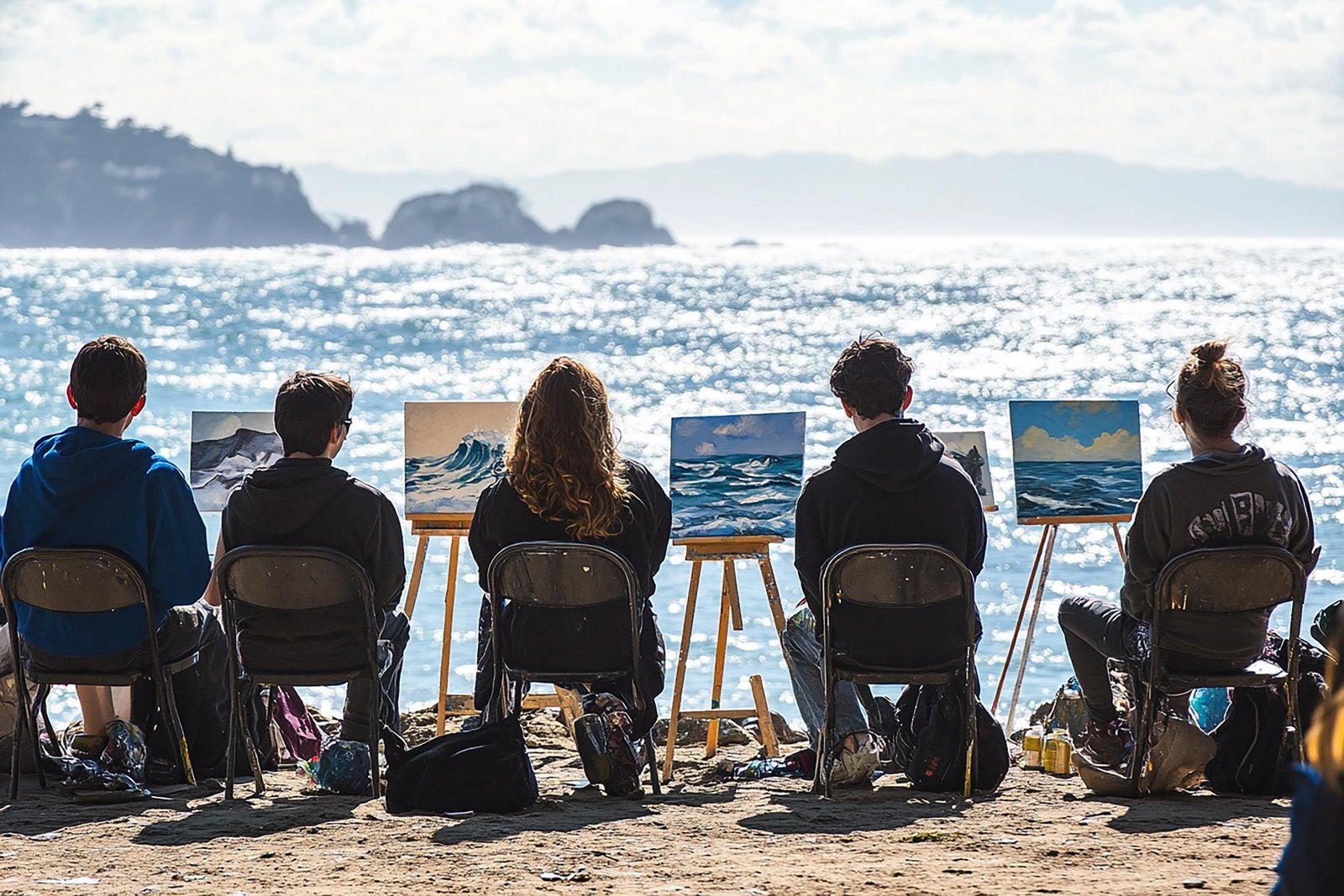 Five people sitting on chairs at the beach facing easels with ocean paintings, looking out at a sparkling ocean with rocky islands in the distance.