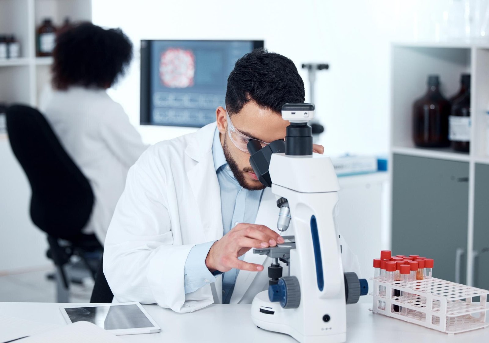 A man in a white lab coat looks through a microscope at a laboratory workstation with test tubes in a rack nearby, while another person in a white coat works at a computer in the background.