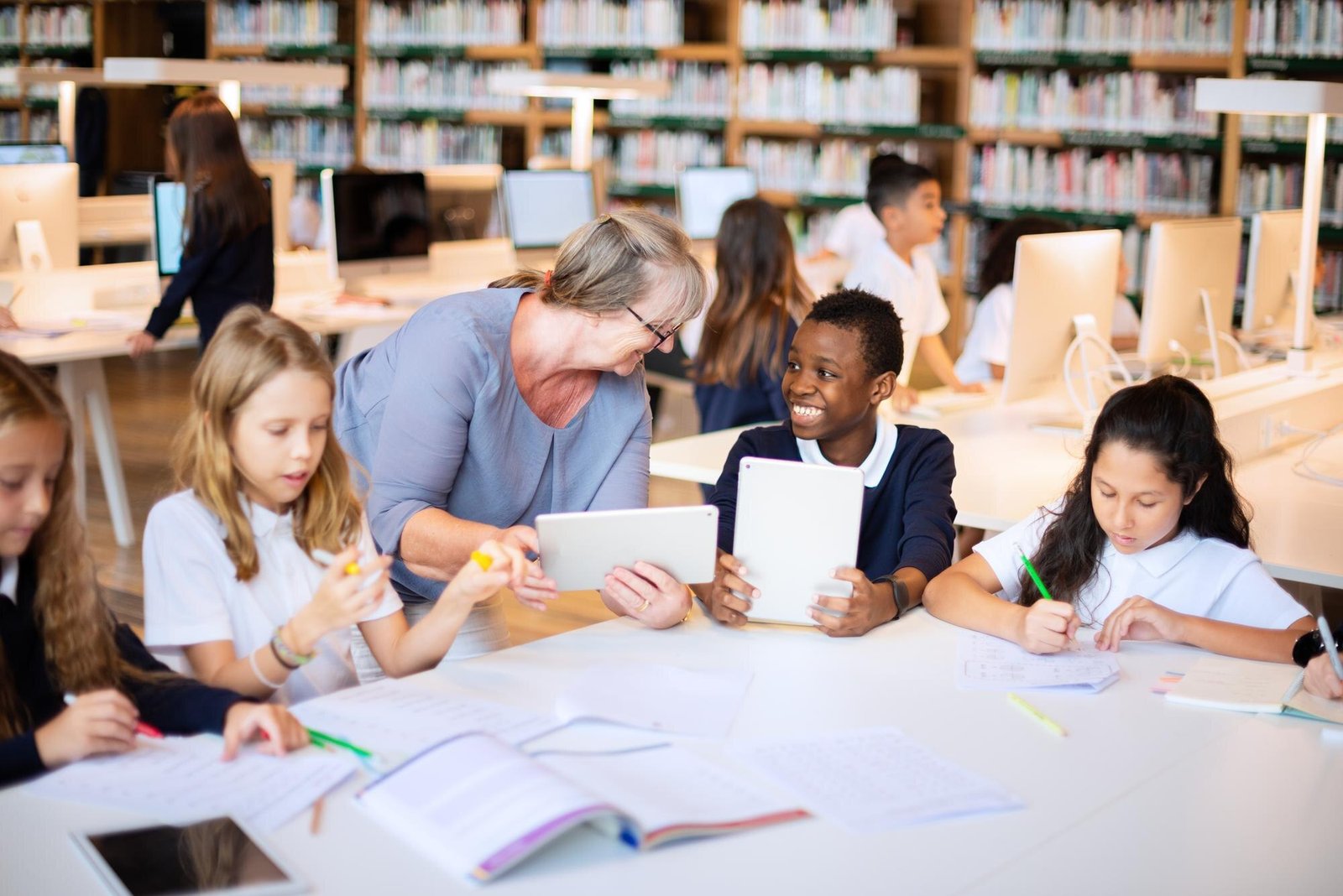 A teacher with gray hair and glasses assists students working with tablets and papers at a table in a library with bookshelves in the background.