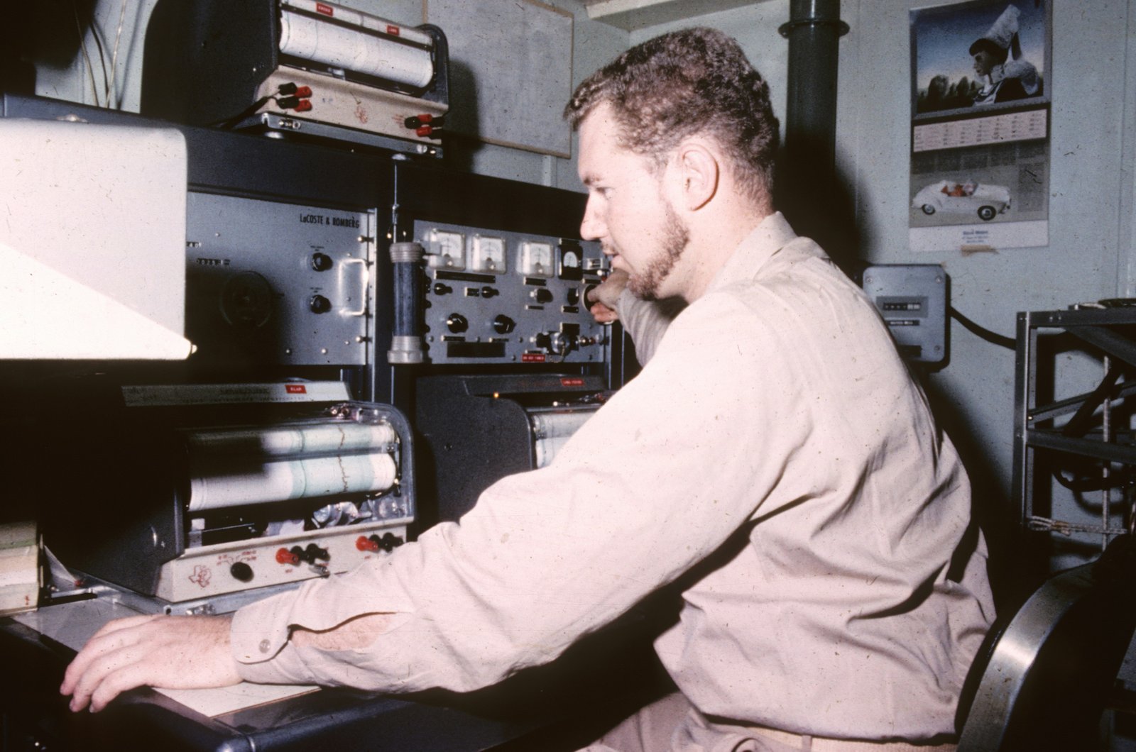A man in a beige/tan colored shirt sits at a desk in a research facility, working at a computer terminal with various electronic equipment and monitoring devices visible in the background, including stacked hardware and wall-mounted photographs.